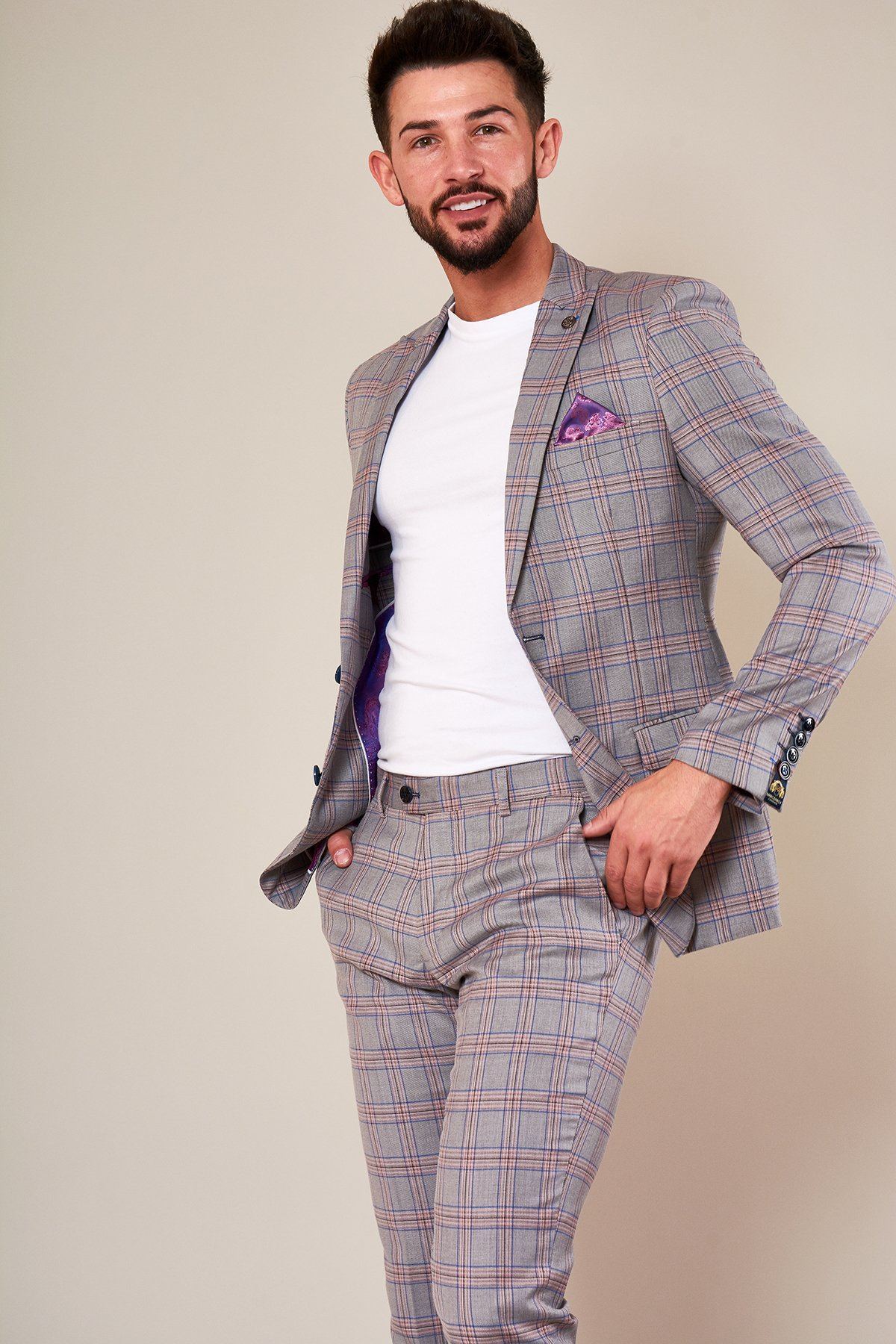 A man with short brown hair and a beard smiles in the Marc Darcy ALVIN - Grey Pink Check Blazer, paired with a purple pocket square and white t-shirt, standing with one hand in his pocket against a neutral background.