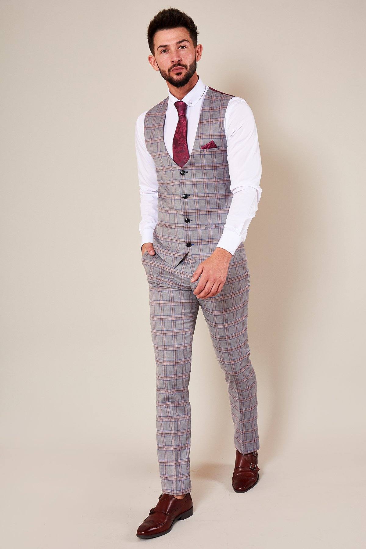A man wears a slim fit grey plaid three-piece suit with a Marc Darcy ALVIN Grey Pink Check Single Breasted Waistcoat, white shirt, maroon tie and pocket square, plus brown dress shoes, standing against a plain beige background with one hand in his pocket.