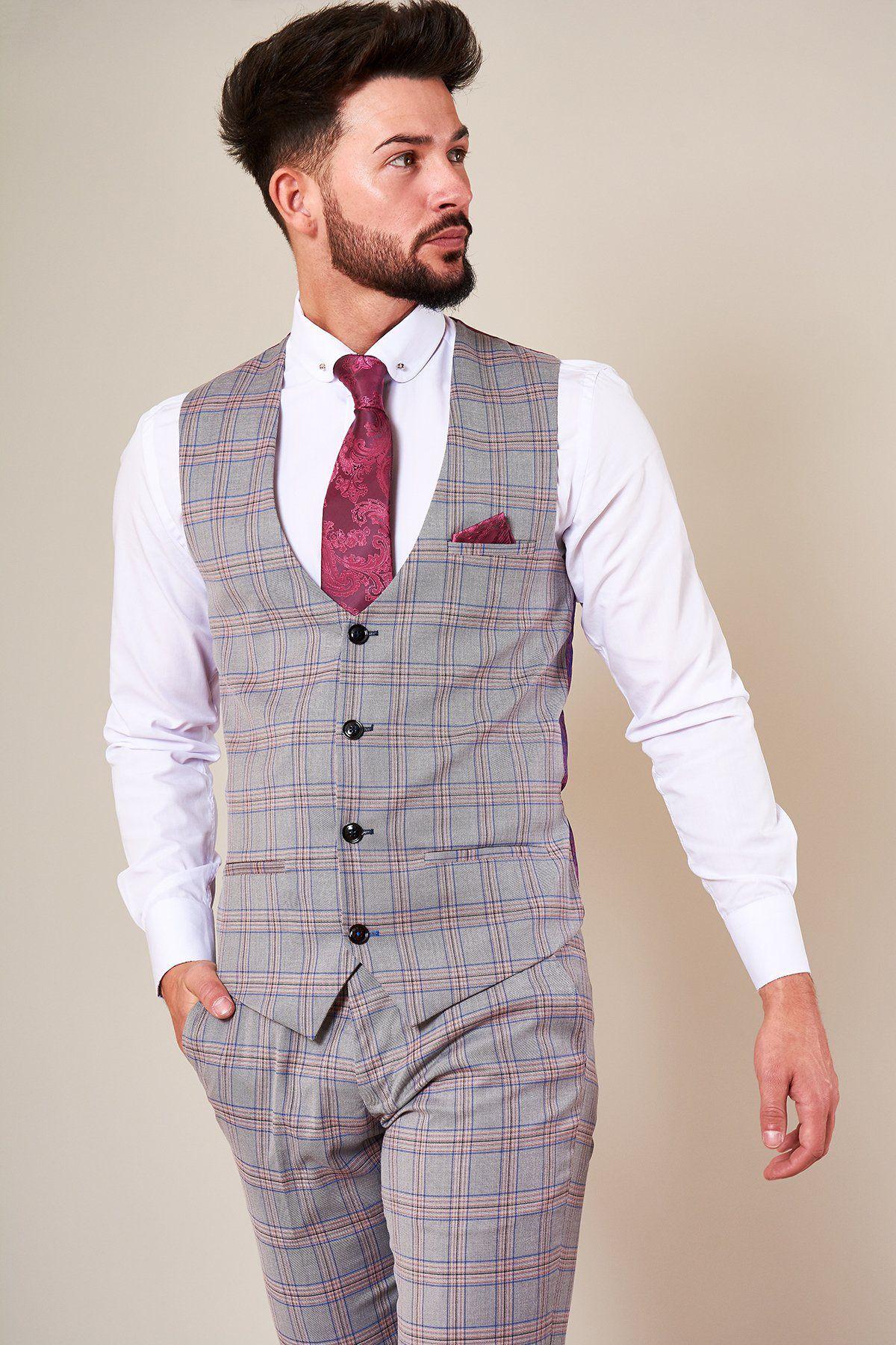 A man with a beard wears the Marc Darcy ALVIN Grey Pink Check Single Breasted Waistcoat with matching trousers, a white dress shirt, and a dark red patterned tie and pocket square. He stands against a plain background, looking to the side with one hand in his pocket.