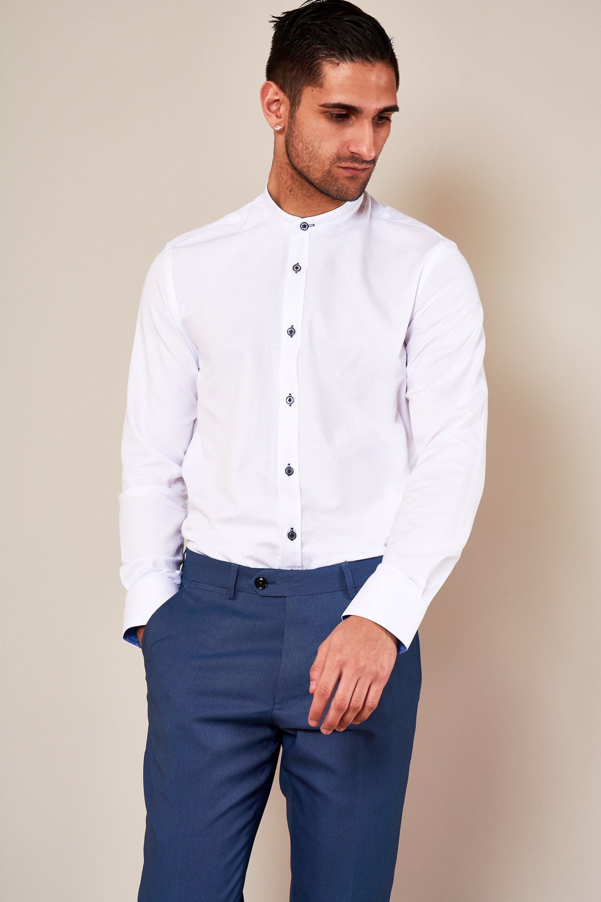 A man with short, dark hair wears the Marc Darcy ARCHIE white grandad collar shirt with navy buttons and blue textured pants, one hand in his pocket.