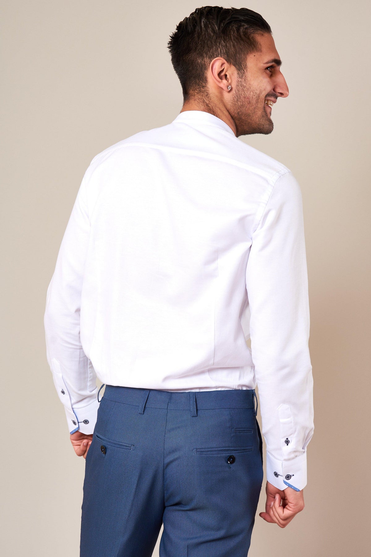 A man wearing the Marc Darcy ARCHIE white grandad collar shirt with navy buttons and blue trousers smiles and looks to the side.