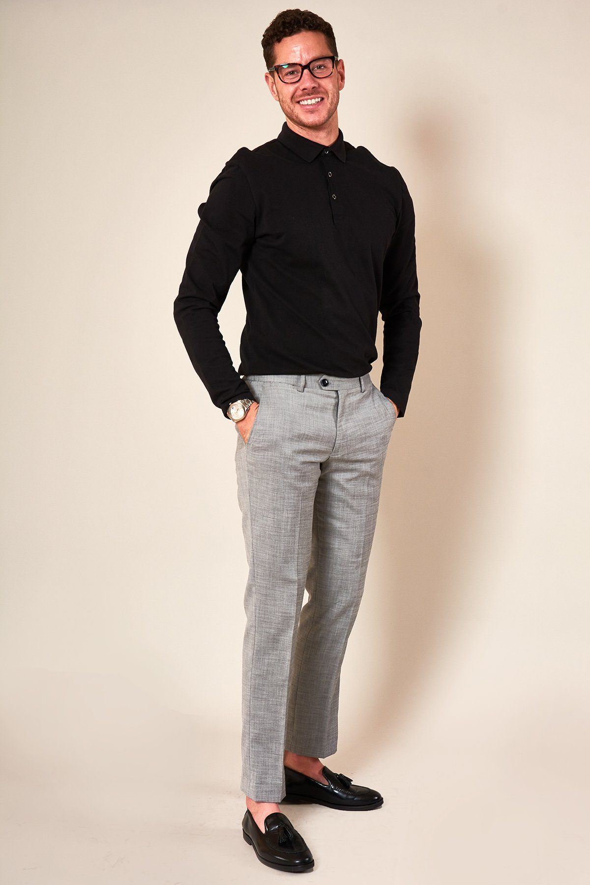 A man in a black long-sleeve polo, marcdarcy BEN - Silver Grey Slim Tailored Trousers, and black loafers smiles at the camera with hands in pockets. Glasses and a watch complete his effortlessly stylish look.