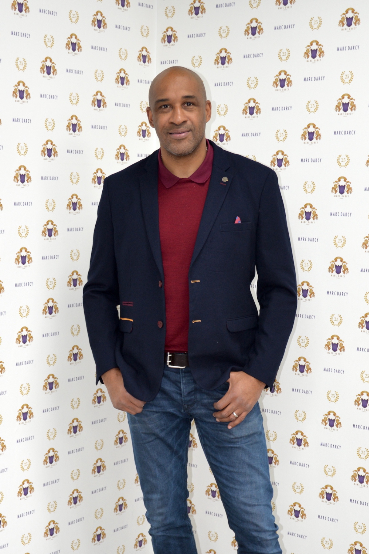 Former Sheffield United footballer Brian Deane smiles against a marcdarcy backdrop, wearing the JD4 Navy Blazer with paisley lining, a red polo shirt, and jeans, one hand in his pocket.