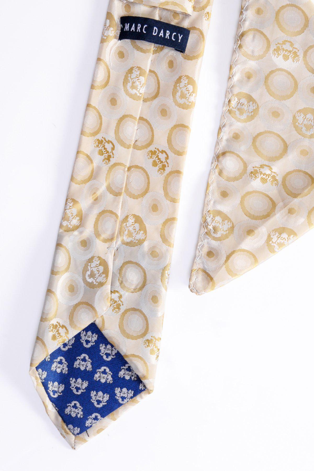 The BUBBLES - Stone Circle Print Tie and Pocket Square Set by Marc Darcy features a handmade beige silk tie with gold circle pattern, complemented by a blue inner tip with white floral design and a sewn-in label, displayed on a white background.