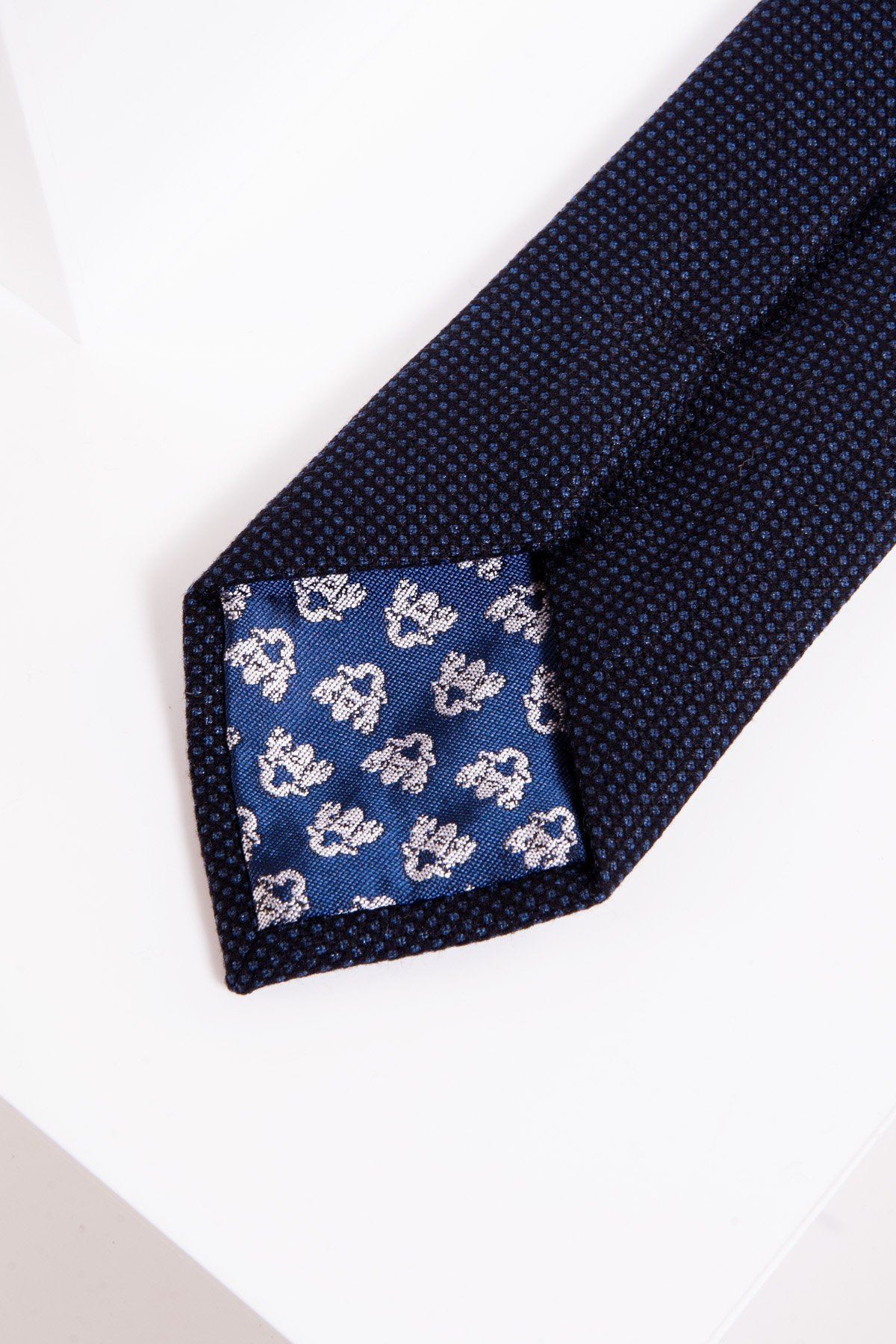 A close-up of the CALLUM - Navy Birdseye Tie by Accessory highlights its birdseye weave and blue and white floral lining, displayed on a white surface.