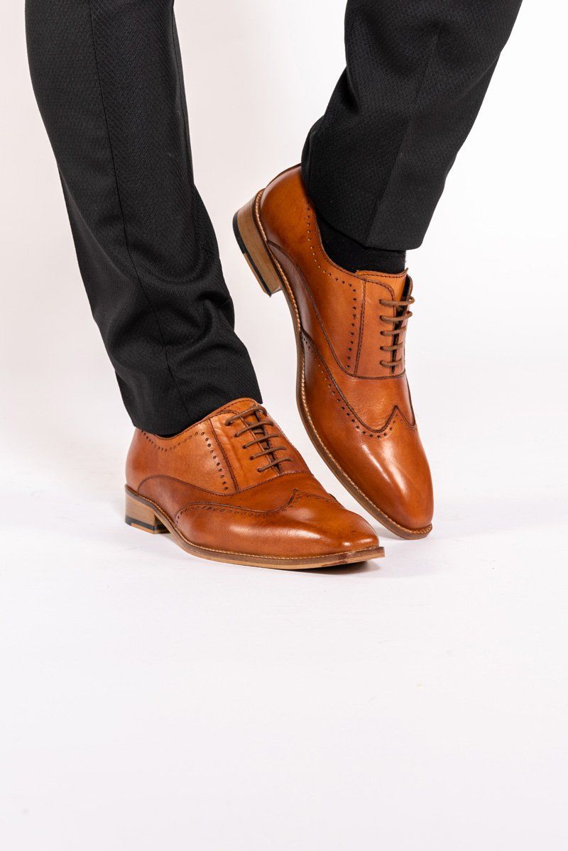 A person stands on a white background wearing black dress pants and the Marc Darcy CARSON – Mid Tan Wingtip Oxford Brogue Shoes, crafted from full grain leather and featuring classic brogue detailing.