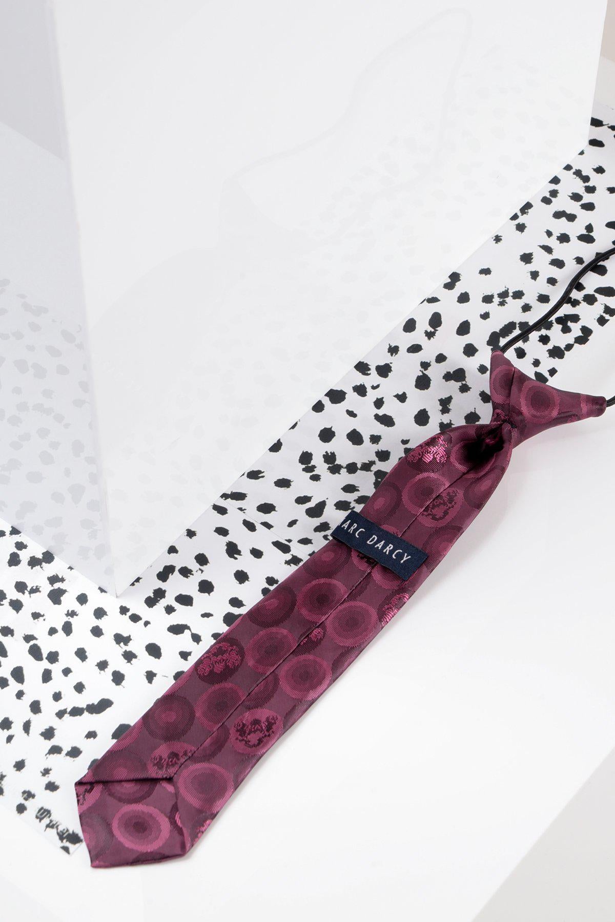 The Marc Darcy Childrens Berry Bubble Circle Print Tie, in maroon pink with a subtle circular pattern, sits on a white surface with black dots—ideal for pairing with a matching adults tie.