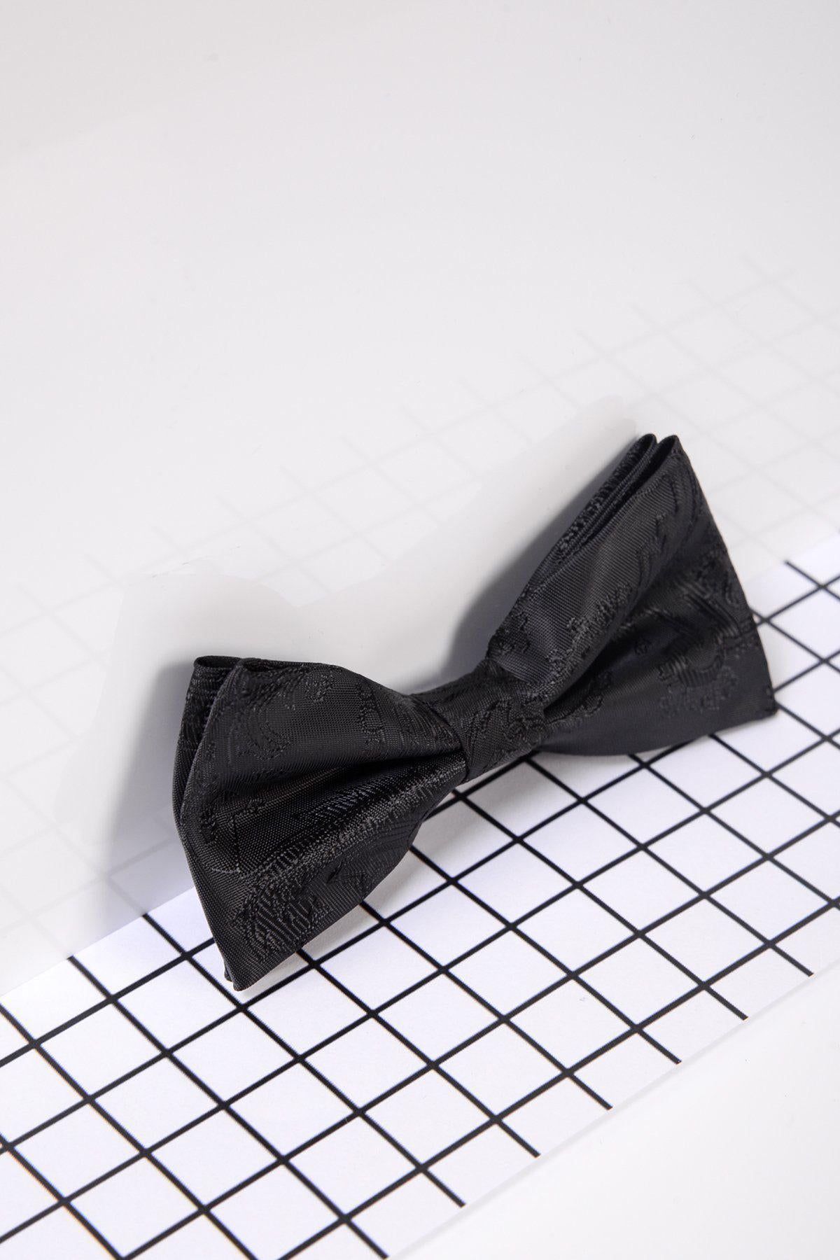 The Marc Darcy Childrens Black Paisley Print Bow Tie, featuring a subtle pattern, is displayed on a black and white grid surface against a plain light background.