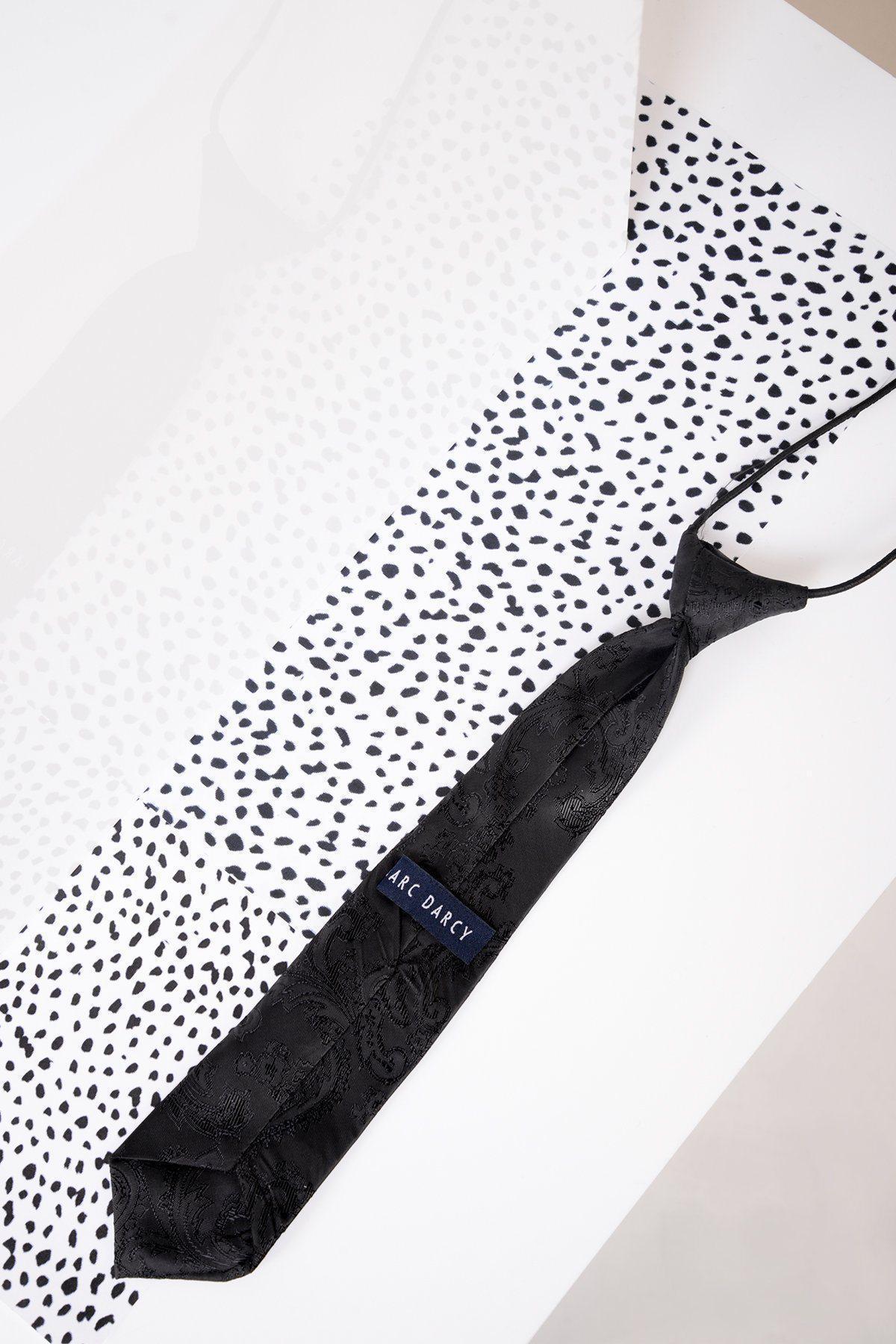 A Childrens Black Paisley Print Tie by Marc Darcy lies on a white, black-speckled surface with a translucent overlay, highlighting its elegant finish.