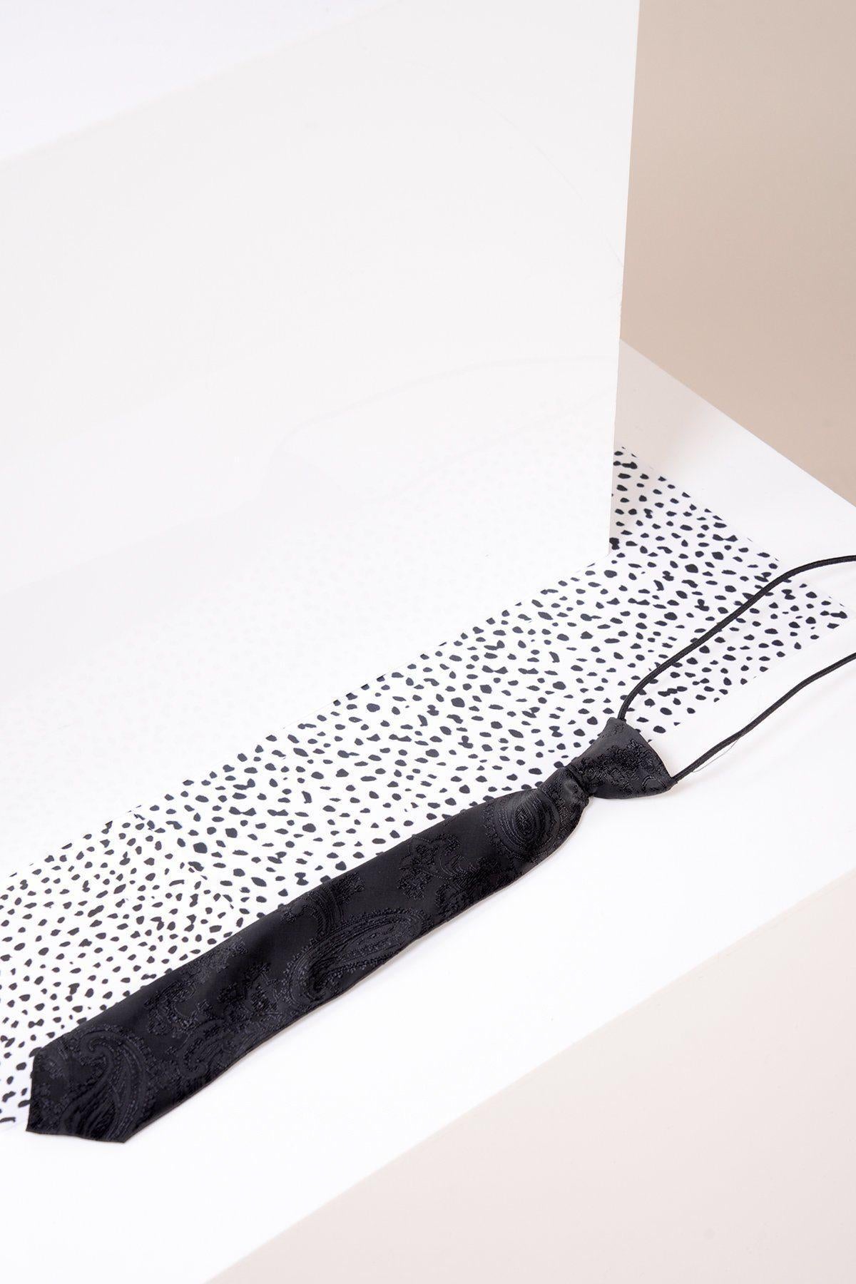 The Marc Darcy Childrens Black Paisley Print Tie, featuring a subtle pattern, rests on a white surface with dotted paper. Geometric beige and white walls in the background emphasize the tie’s refined elegance.