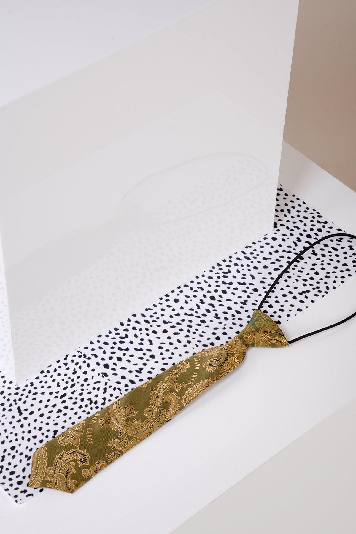 A Children’s Gold Tan Paisley Print Tie by Marc Darcy, featuring a black adjustable elastic neck strap, rests on a speckled white surface and is partially reflected in an upright clear acrylic sheet.