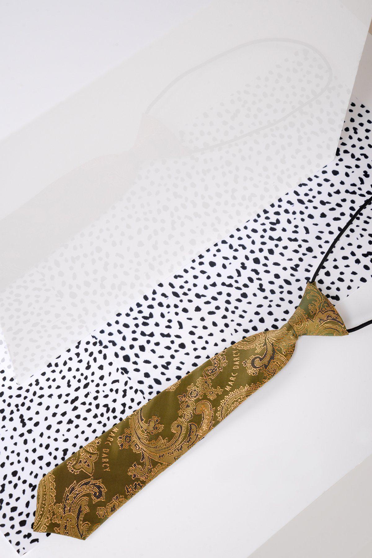 A Marc Darcy Children's Gold Tan Paisley Print Tie rests on a white surface with black irregular dots, beside a reflective panel that reveals the tie’s subtle reflection.