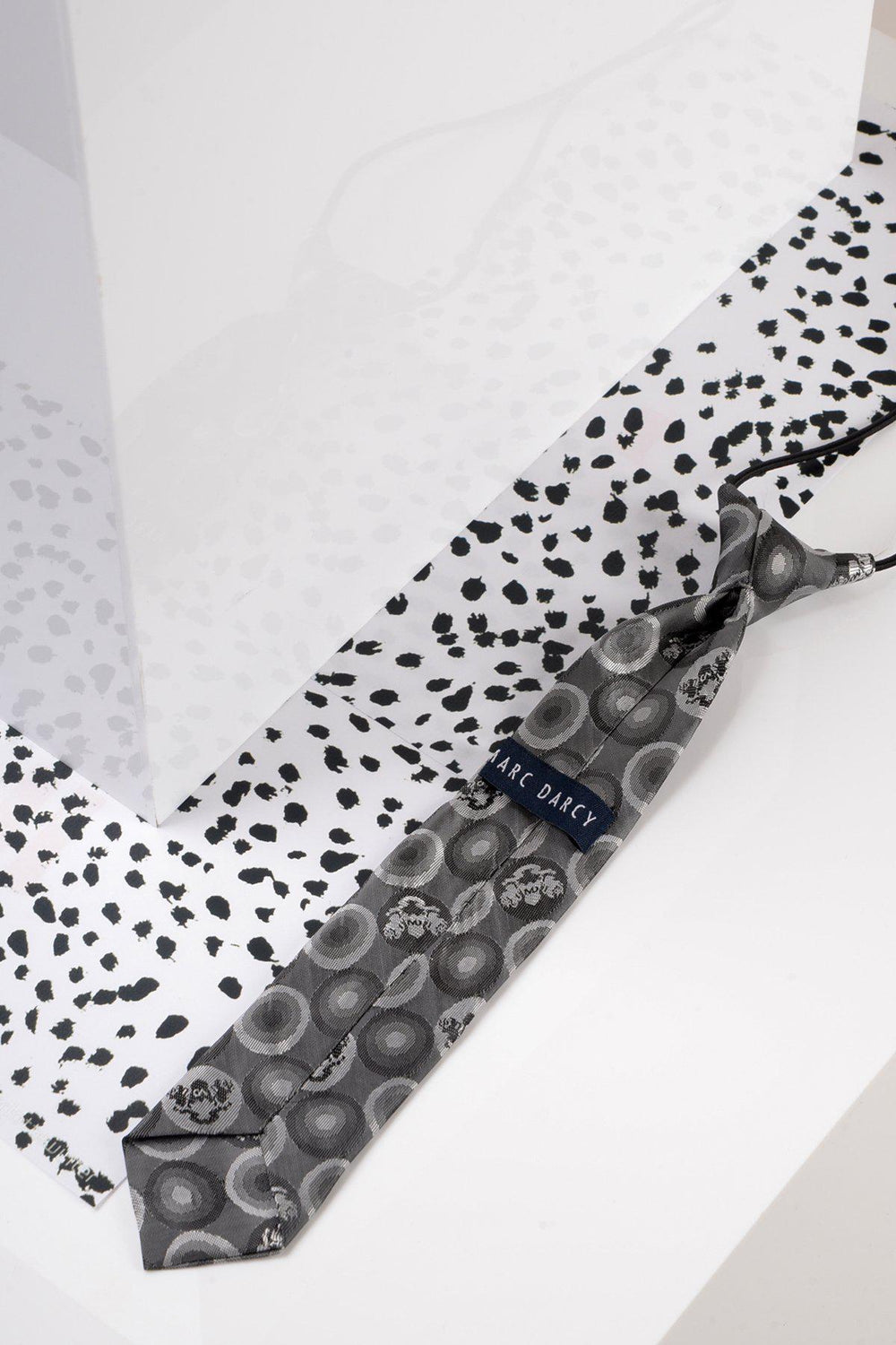 A Childrens Grey Bubble Circle Print Tie by Marc Darcy lies on a white surface with black dot patterns, next to a glossy white box. The tie features circular designs and is ideal for matching with an adults version.