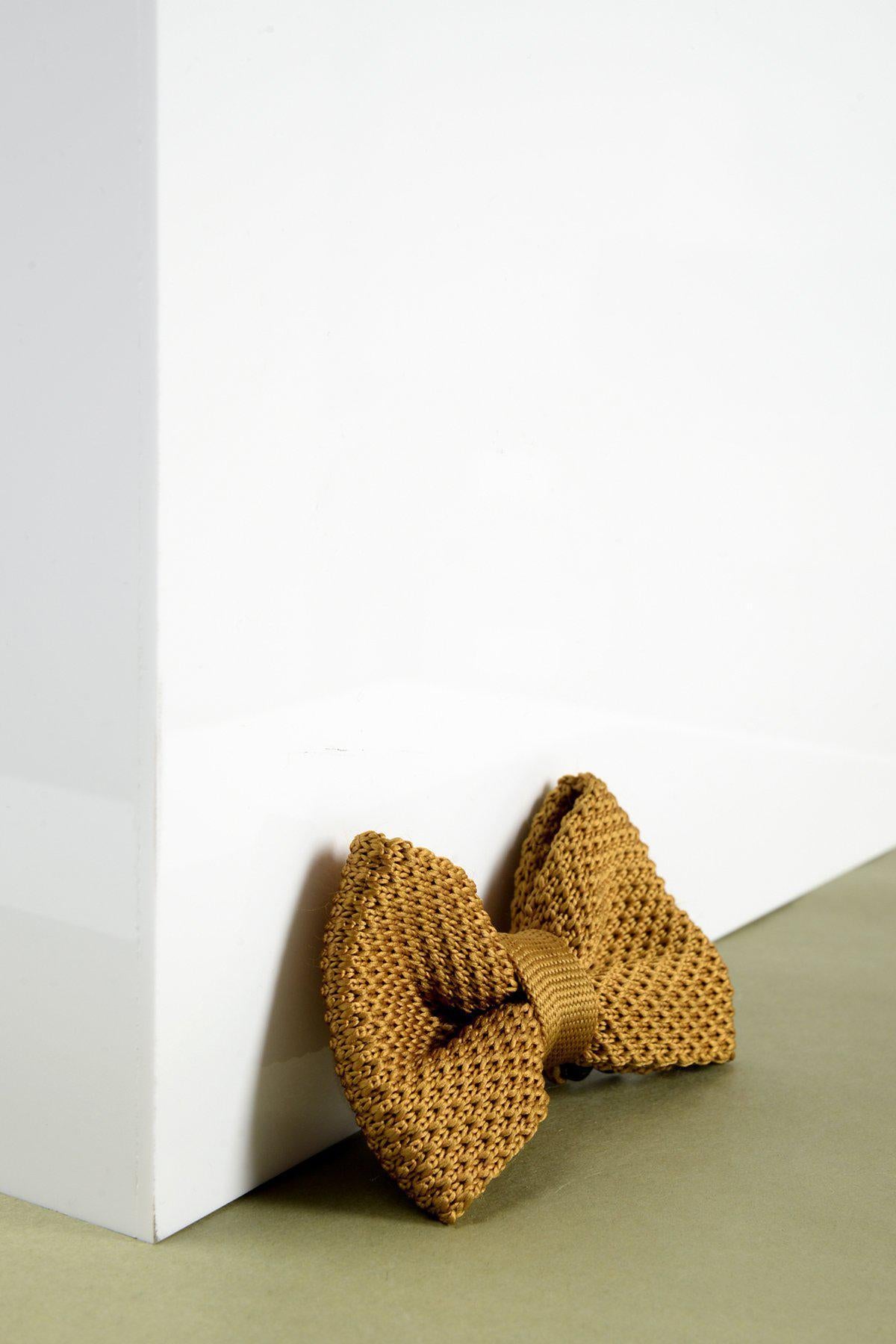 A close-up of the Marc Darcy Children's Knitted Bow Tie In Gold, an adjustable bow tie featuring a rich golden-brown knit, perfect for stylish kids and shown leaning against a white corner.