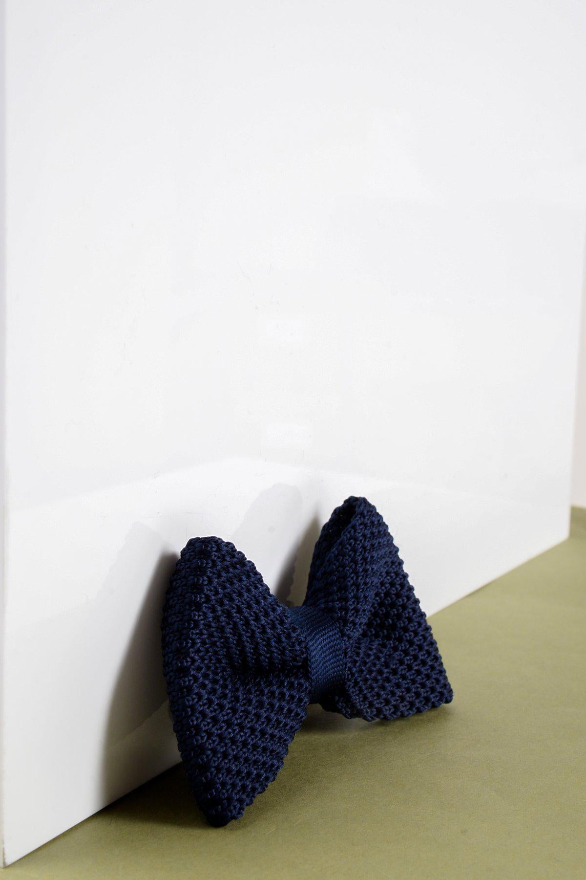 The Marc Darcy Children's Knitted Bow Tie in Navy stands upright against a white surface on a light green background—a stylish accessory ideal for boys' suits at special occasions.