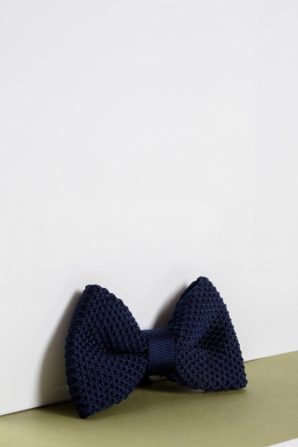 The Marc Darcy Childrens Knitted Bow Tie in Navy is displayed on a white background with a light olive-green surface, featuring an adjustable strap for comfort.