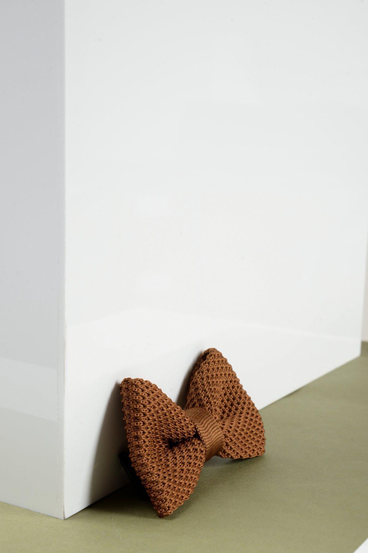 The Marc Darcy Children's Knitted Bow Tie in Rust rests at the base of a large white rectangular object on a muted green surface, ideal for formal kids' accessories.
