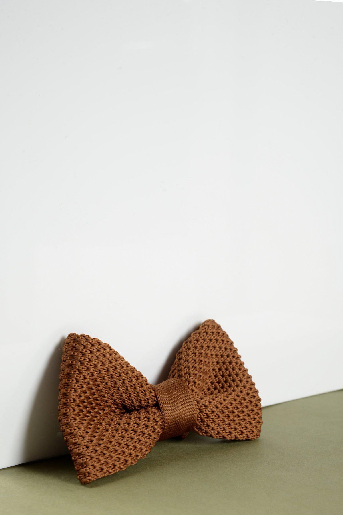 The Marc Darcy Childrens Knitted Bow Tie in Rust, featuring a modern knitted texture, is displayed upright and neatly tied against a white surface with a green base.