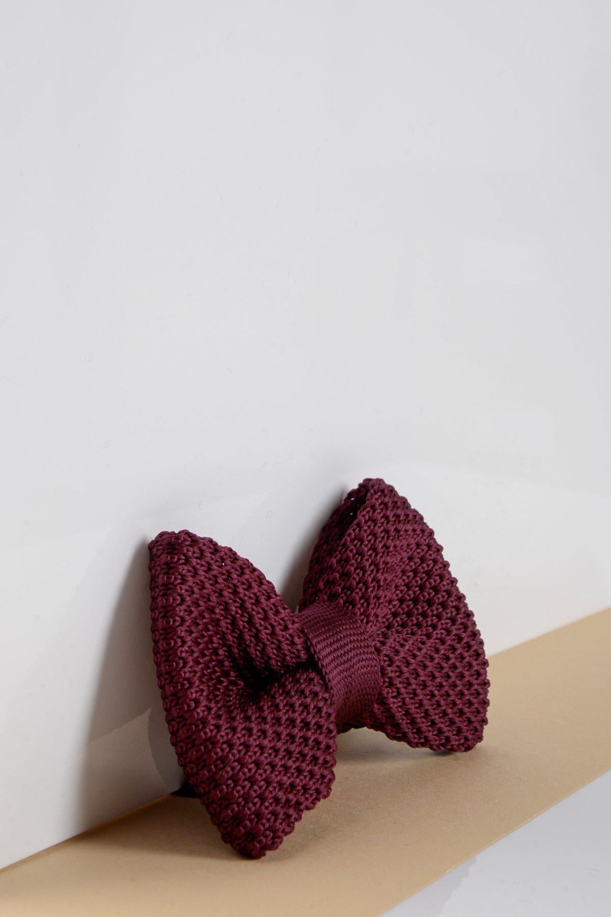 The Marc Darcy Childrens Knitted Bow Tie in Wine stands upright on a white surface, set against a beige backdrop at the bottom.