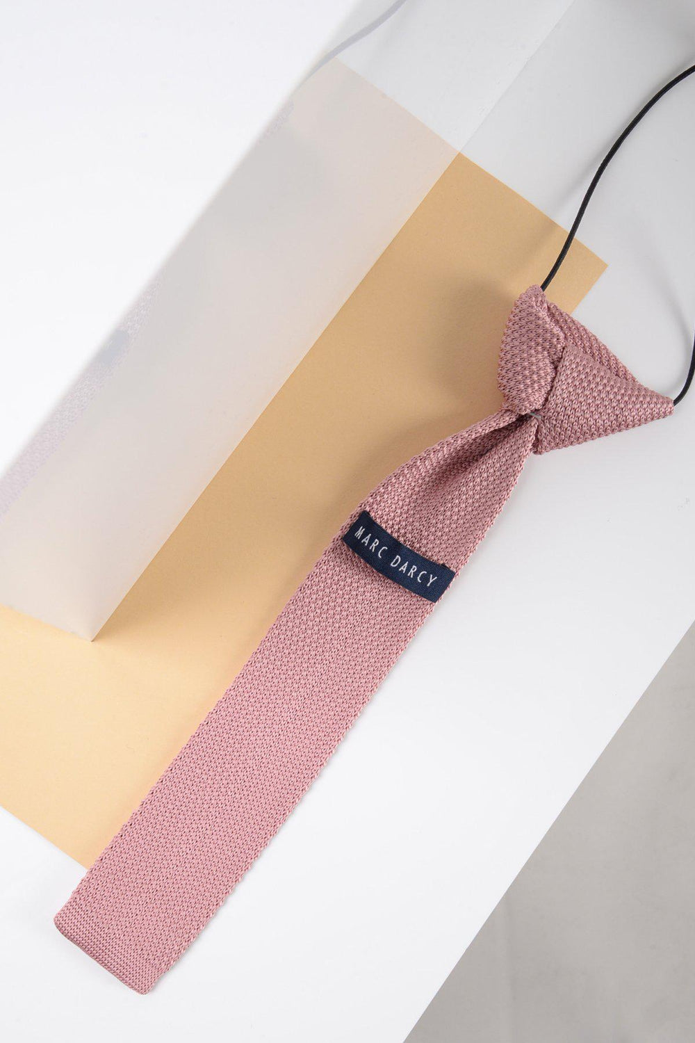 The Children's Knitted Tie In Blush Pink by Marc Darcy features a textured finish and adjustable neck strap—ideal for formal occasions. Photographed on a beige and white geometric background, it adds charm to any outfit.