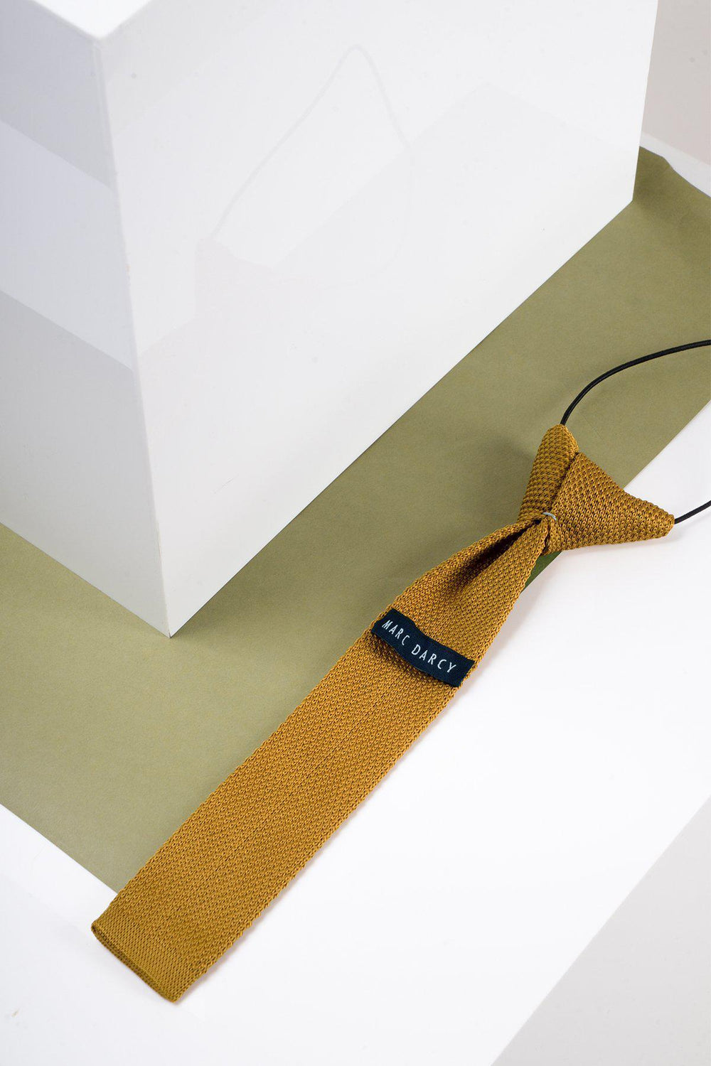 A Marc Darcy Childrens Knitted Tie in gold with an adjustable neck strap is displayed on a light olive-green surface beside a white geometric object.