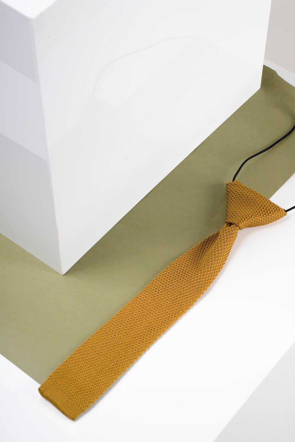 The Marc Darcy Children's Knitted Tie in Gold is displayed diagonally on a white and olive green surface beside a white geometric block.