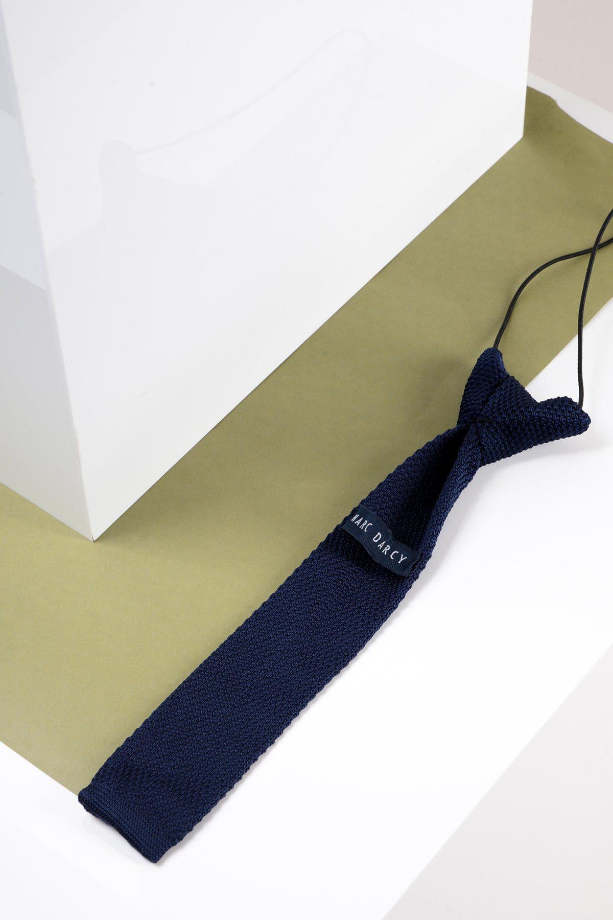 A Childrens Knitted Tie in Navy by Marc Darcy with an adjustable strap lies on an olive green and white geometric surface next to a white rectangular object.
