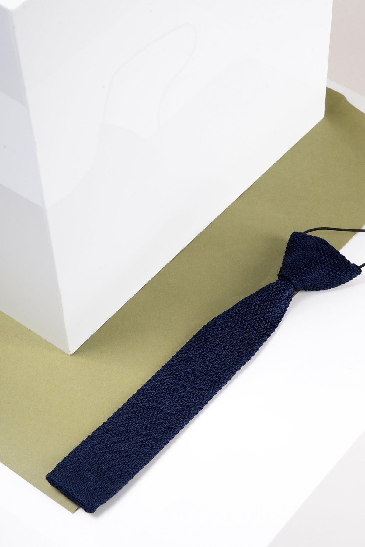 The Marc Darcy Children's Knitted Tie in Navy, featuring a textured finish, is displayed on a light green surface beside a white geometric structure.