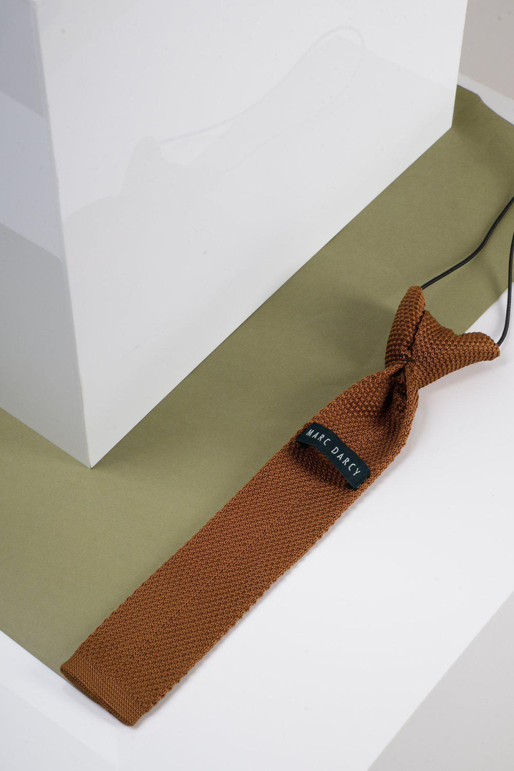 The Children's Knitted Tie in Rust by Marc Darcy is displayed on an olive green surface next to a white geometric object, set against a white background—ideal as a formal boys' accessory.