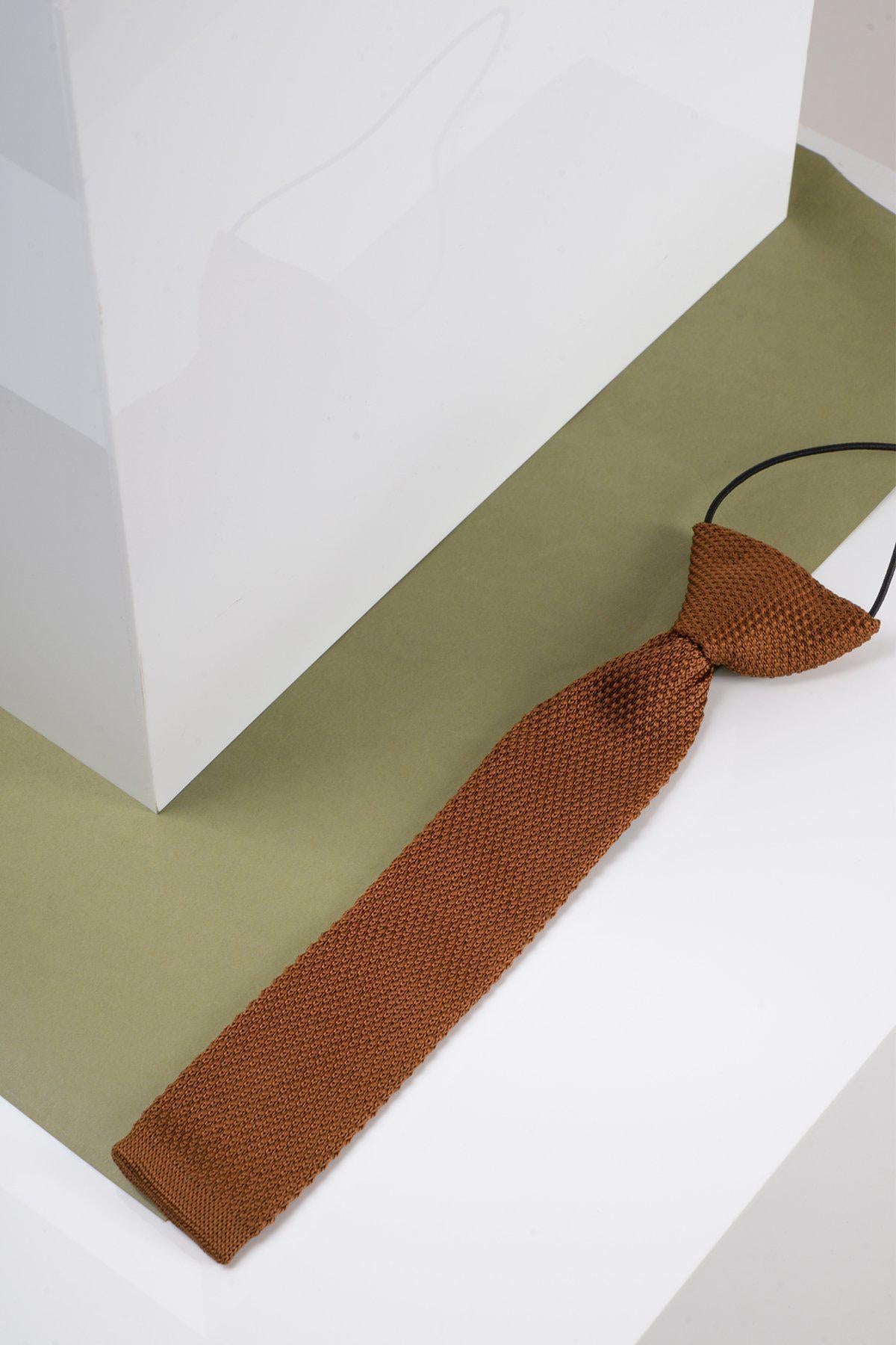 The Marc Darcy Children's Knitted Tie in Rust is displayed on a green and white geometric surface beside a white rectangular object, making it a standout formal accessory for kids.