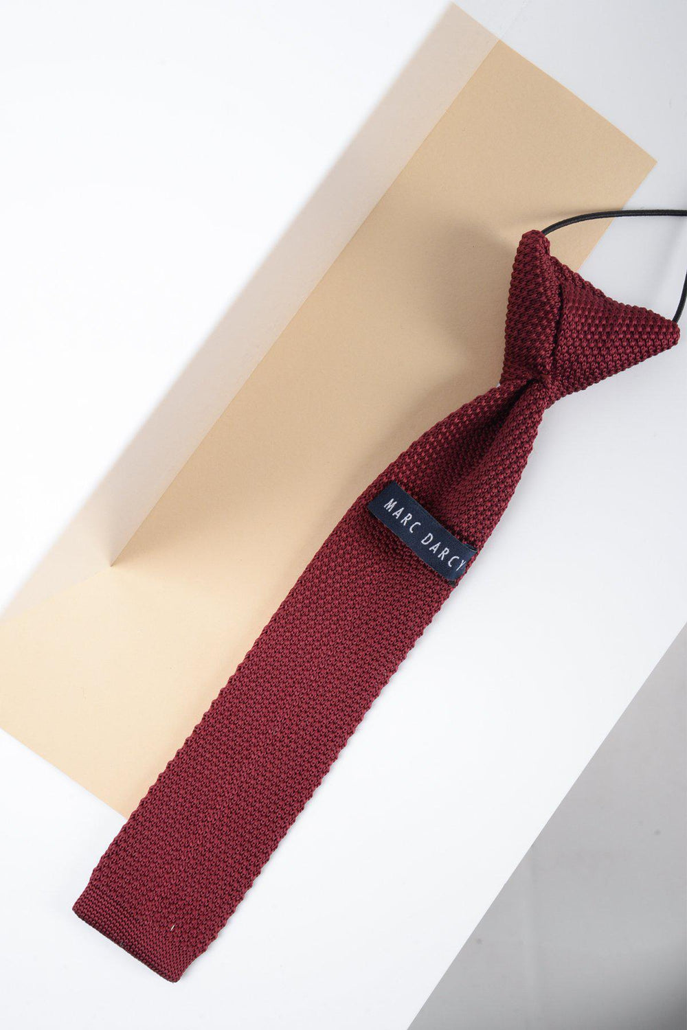 A Children’s Knitted Tie in wine by Marc Darcy is placed diagonally on a beige envelope against a white surface.