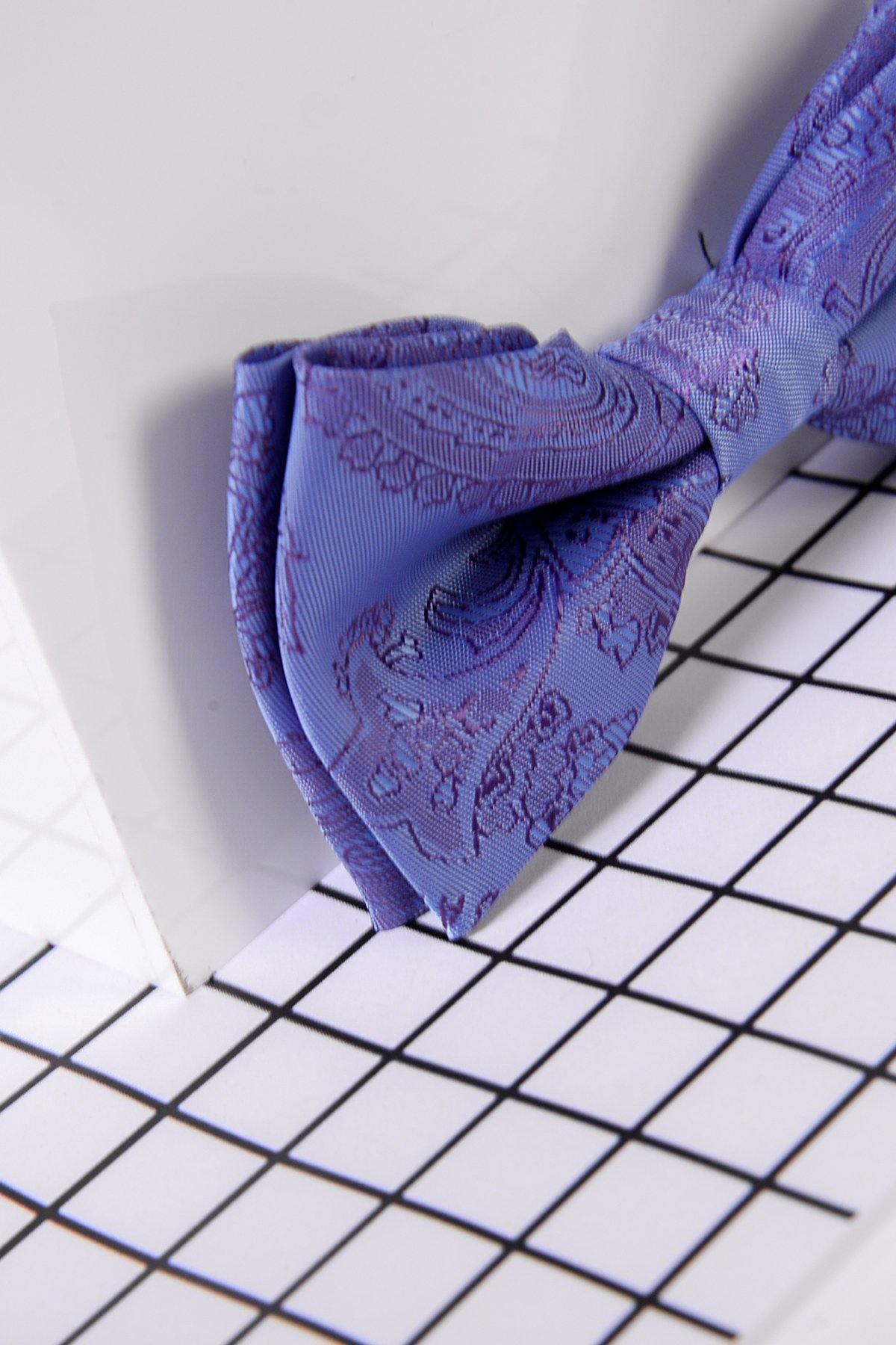 The Marc Darcy Childrens Lilac Paisley Print Bow Tie, featuring an adjustable neck strap, is shown close-up on a white surface with a black grid pattern, partially resting against a glossy white background.