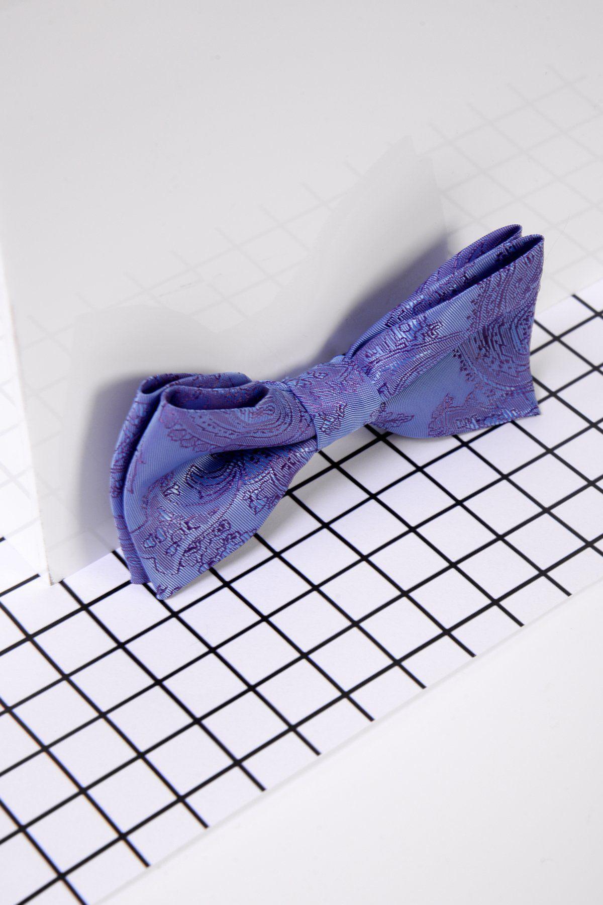 The Marc Darcy Children's Lilac Paisley Print Bow Tie rests on a black-and-white grid surface by a white wall, featuring an adjustable neck strap ideal for boys' formal occasions.