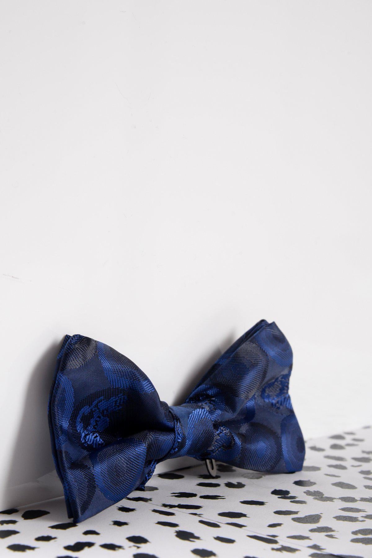 The Marc Darcy Childrens Navy Bubble Circle Print Bow Tie, featuring a subtle circular pattern and adjustable neck strap, is displayed on a white surface with black speckled spots against a white wall.