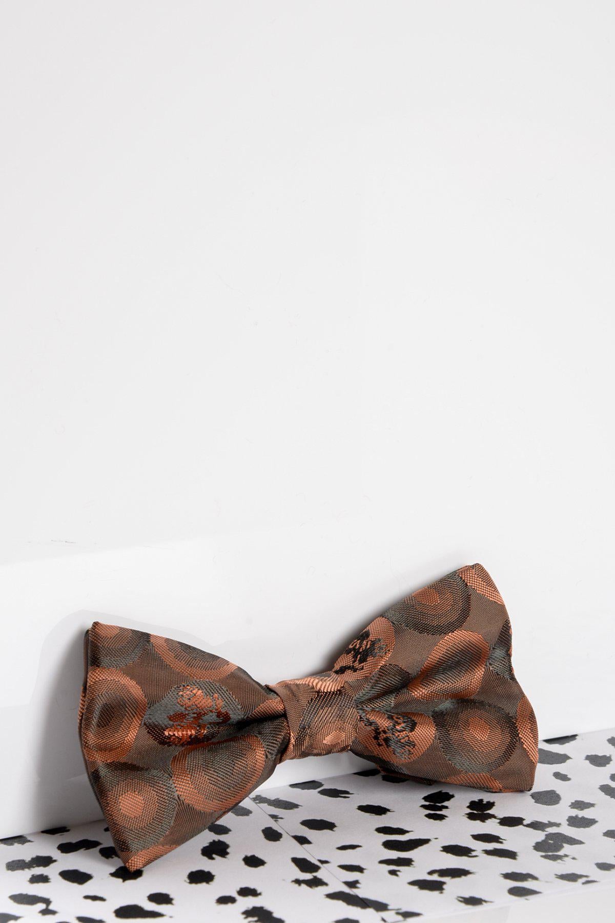 The Marc Darcy Children's Orange Brown Bubble Circle Print Bow Tie, featuring an adjustable neck strap, sits on a white surface with black speckles near the edge. The background is plain and light-colored.