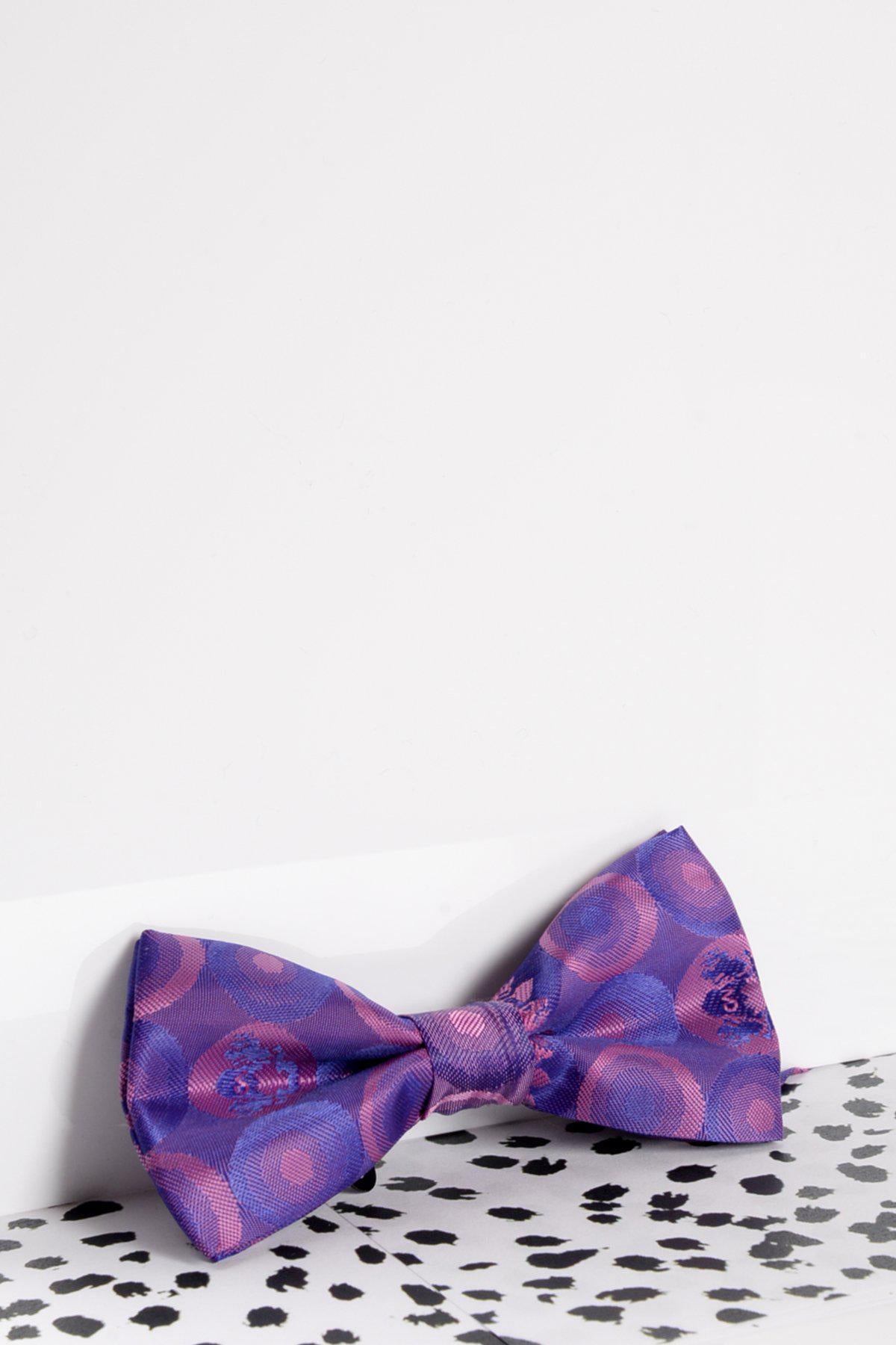 The Marc Darcy Children's Purple Bubble Circle Print Bow Tie rests on a white surface beside an animal print background, featuring an adjustable strap—perfect for boys' partywear.