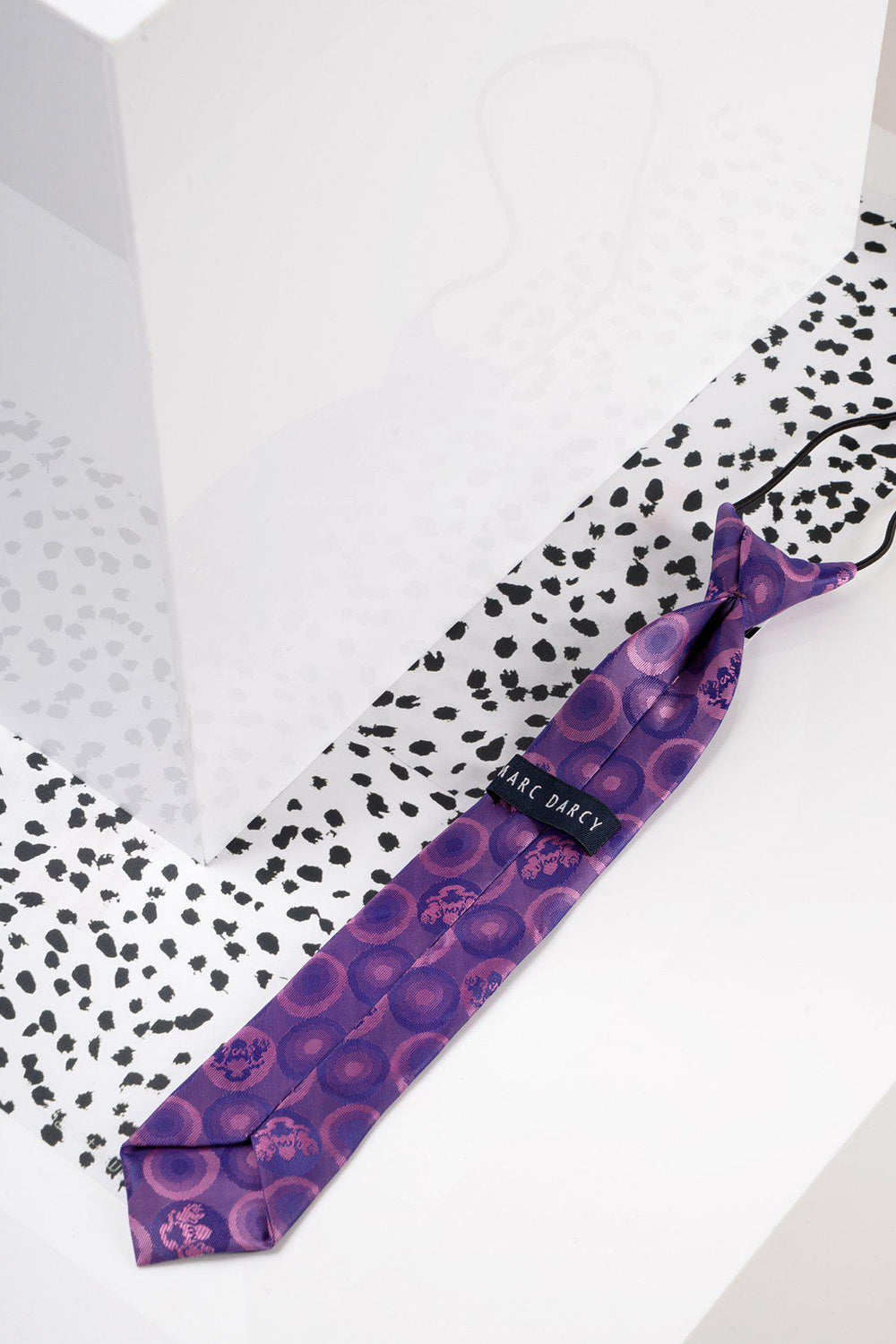 The Marc Darcy Children's Purple Bubble Circle Print Tie rests on a white surface with black spots beside a glossy geometric object—ideal for pairing with boys’ partywear or swapping for an adjustable silk bow tie.
