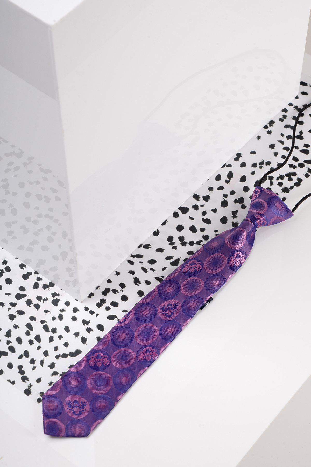 The Marc Darcy Children's Purple Bubble Circle Print Tie, featuring a vibrant circular pattern, rests on a white surface with black dots beside a large white geometric block. Ideal for boys' partywear.