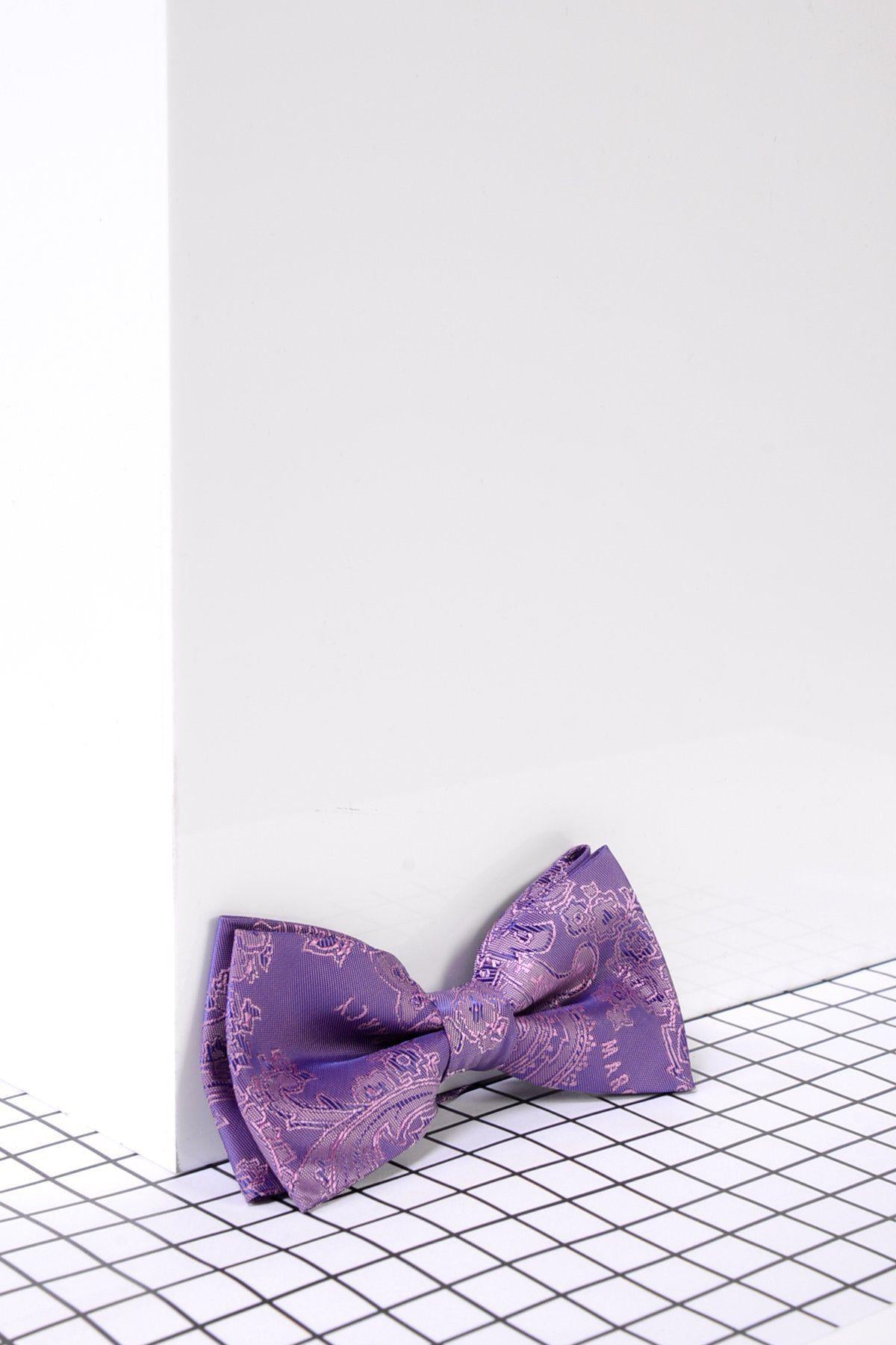 The Marc Darcy Children's Purple Paisley Print Bow Tie, a stylish formal accessory with an adjustable neck strap, is displayed at the base of a white wall on a black and white grid-patterned surface.