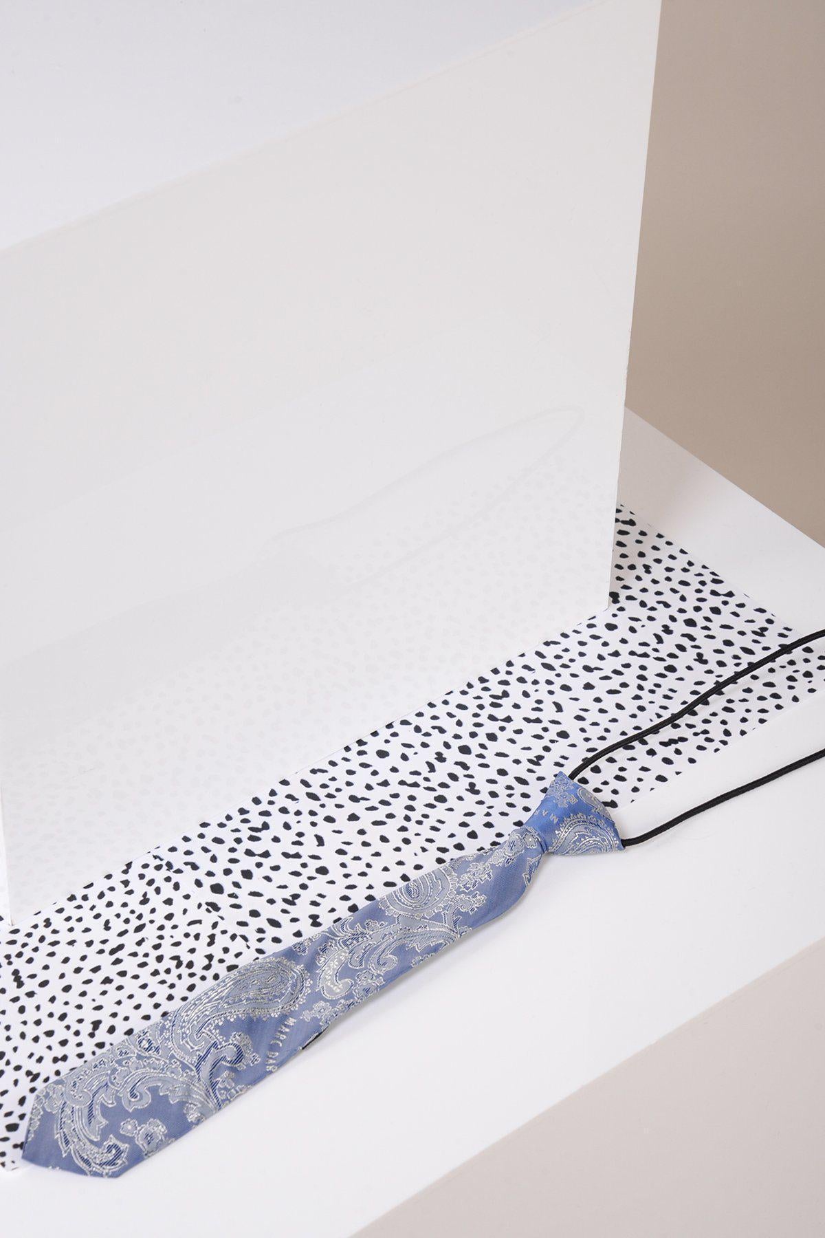 The Marc Darcy Children's Sky Blue Paisley Print Tie, with silver designs, rests on a white surface and black-and-white spotted fabric—an ideal formal accessory for any page boy suit.
