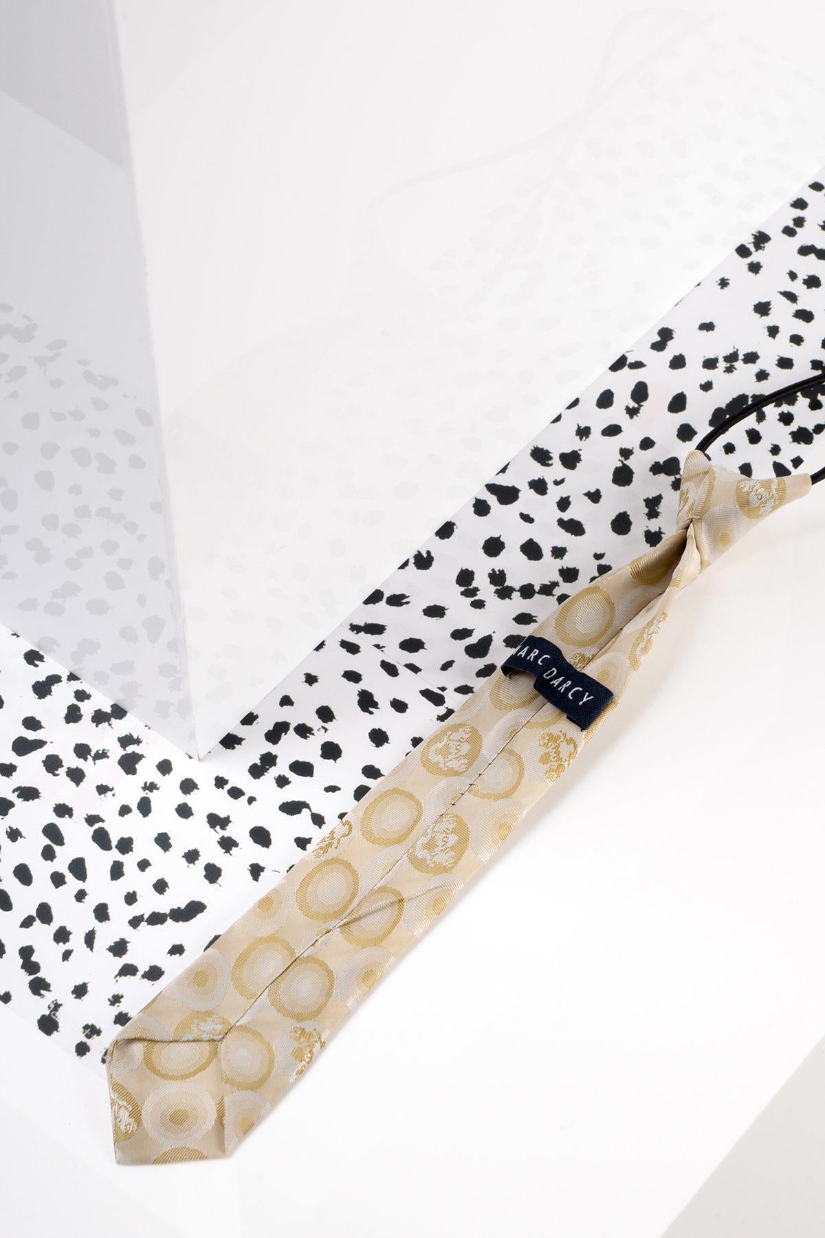 The Children's Stone Bubble Circle Print Tie by Marc Darcy features a beige base with light yellow circles and a visible brand label, shown on a white surface, making it an ideal formal accessory for kids. A black and white speckled pattern is in the background.