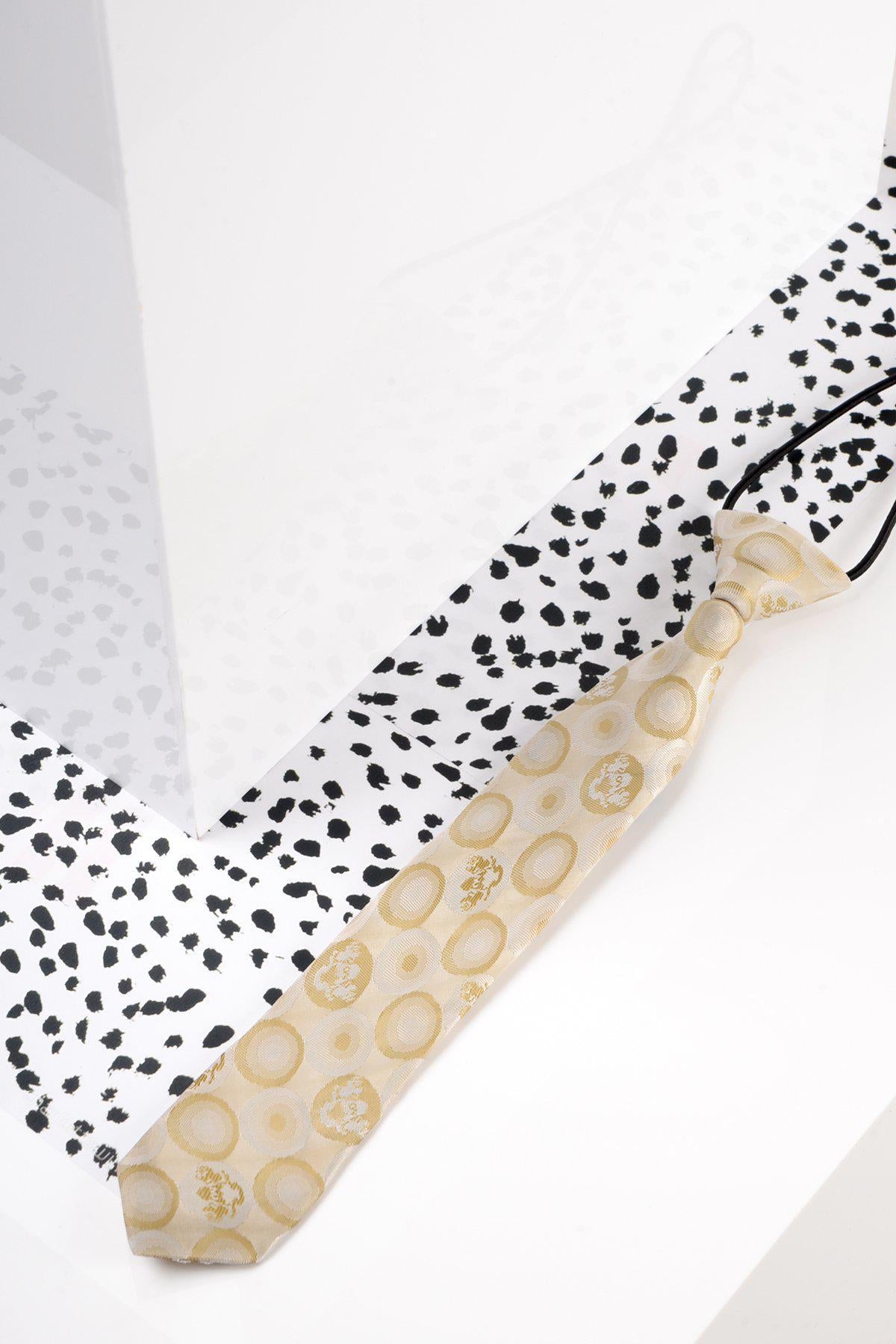 A Marc Darcy Children’s Stone Bubble Circle Print Tie rests on a white surface with black spots, beside a glossy white wall that reflects light and casts a faint shadow.
