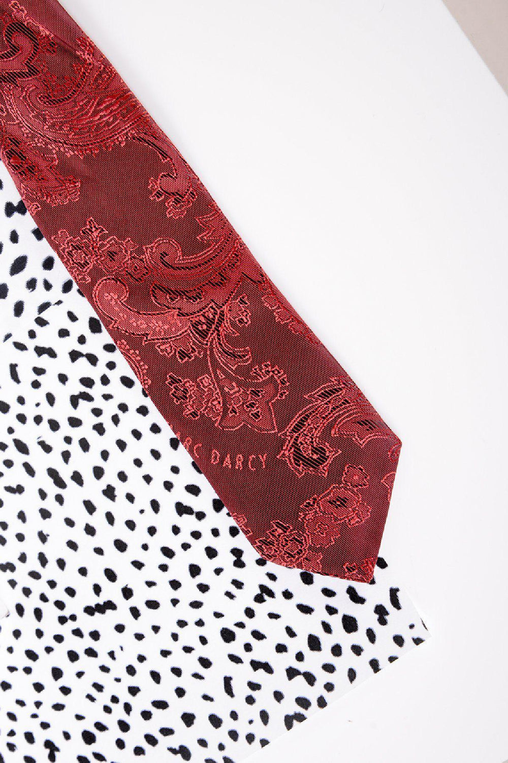 The Childrens Wine Paisley Print Tie by Marc Darcy rests on a white surface with black spots resembling a Dalmatian print. Perfect for formal outfits or a page boys suit, it features Marc Darcy subtly embroidered near the tip.