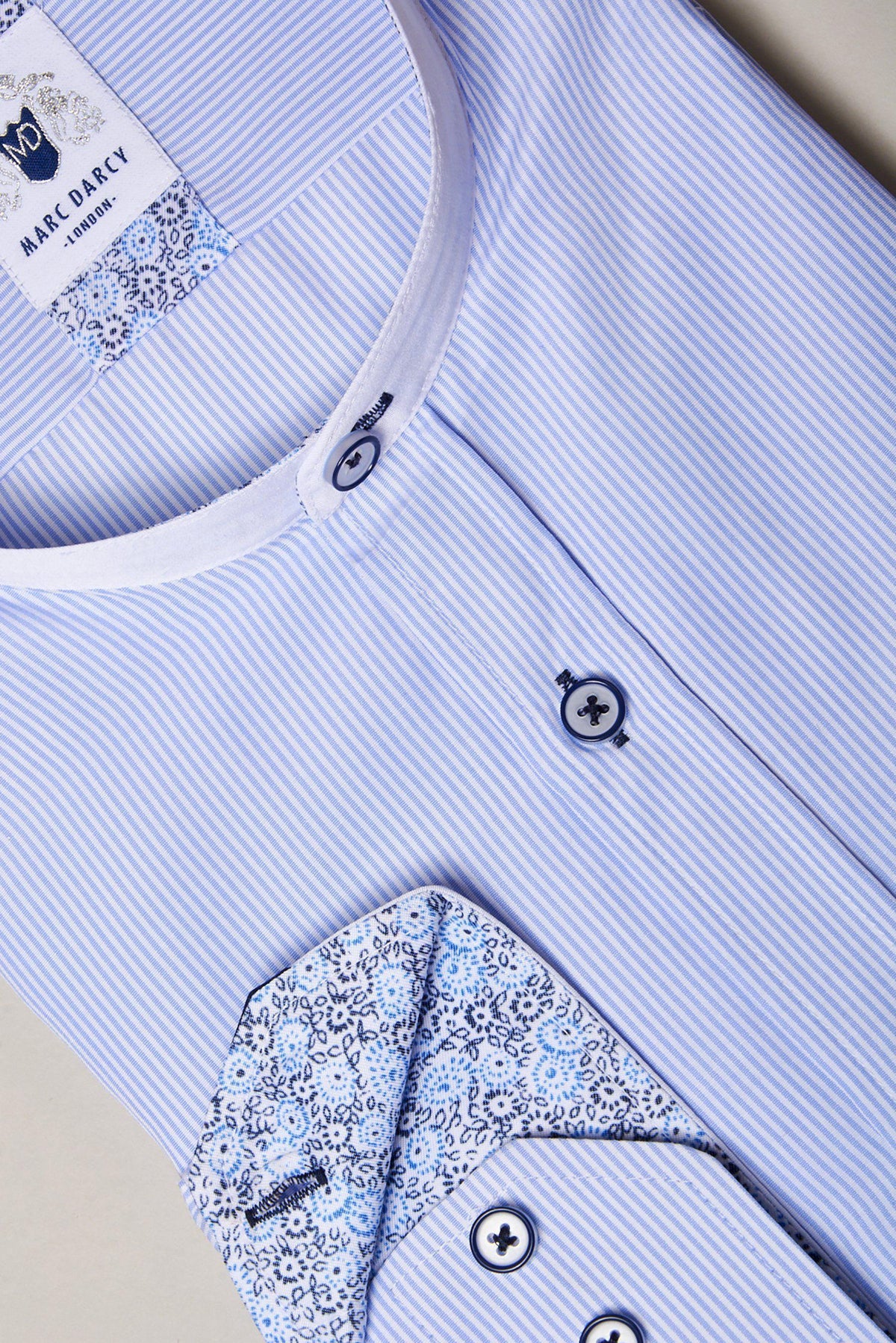 A close-up of the Marc Darcy COOPER Grandad Collar Sky Blue Stripe Shirt, featuring light blue and white stripes, a rounded grandad collar, floral inner lining on the collar and cuffs, and dark buttons.