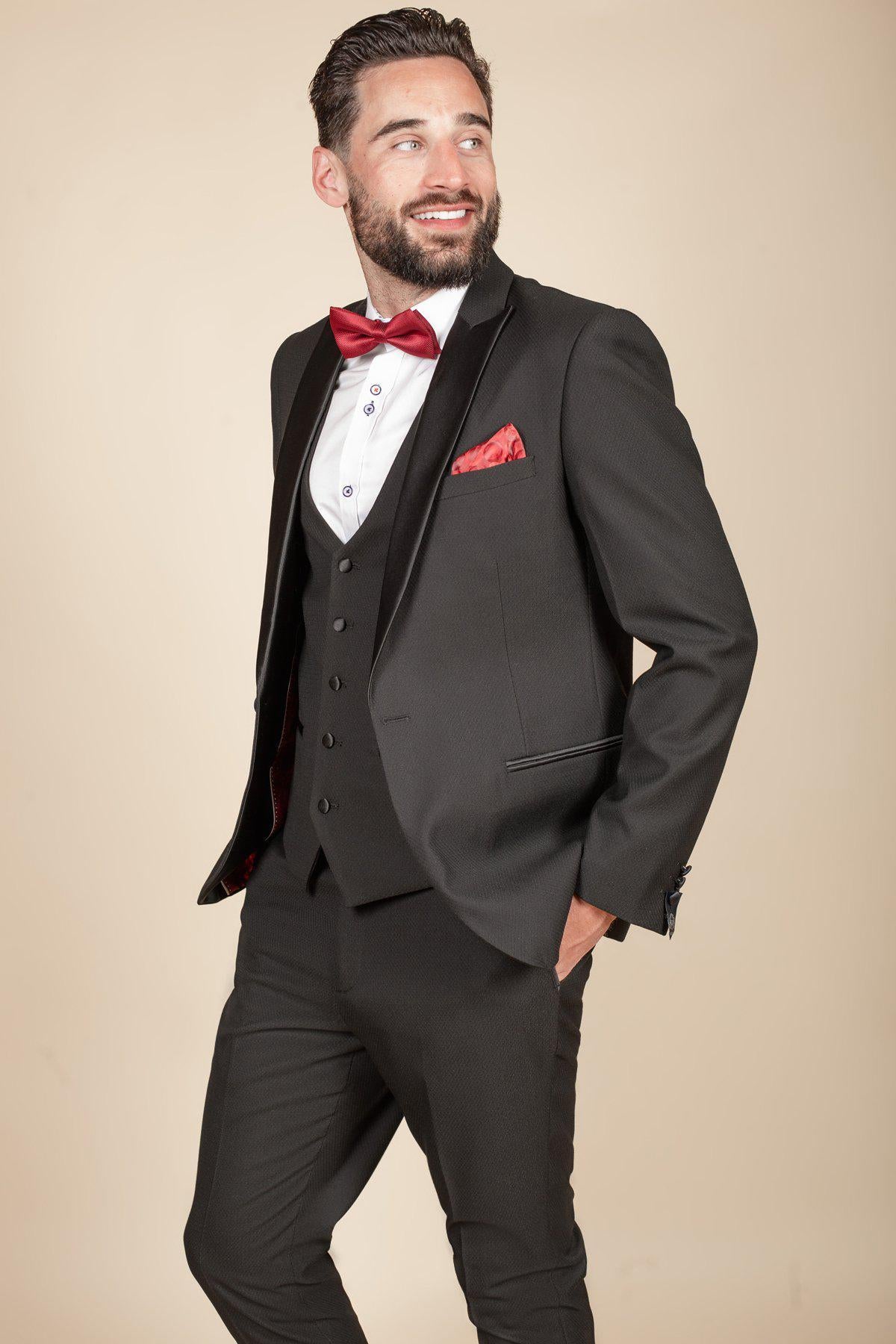Former Crystal Palace footballer Clinton Morrison wears the marcdarcy DALTON Black Tux Lapel Suit with a red bow tie and pocket square, smiling with one hand in his pocket against a beige background.