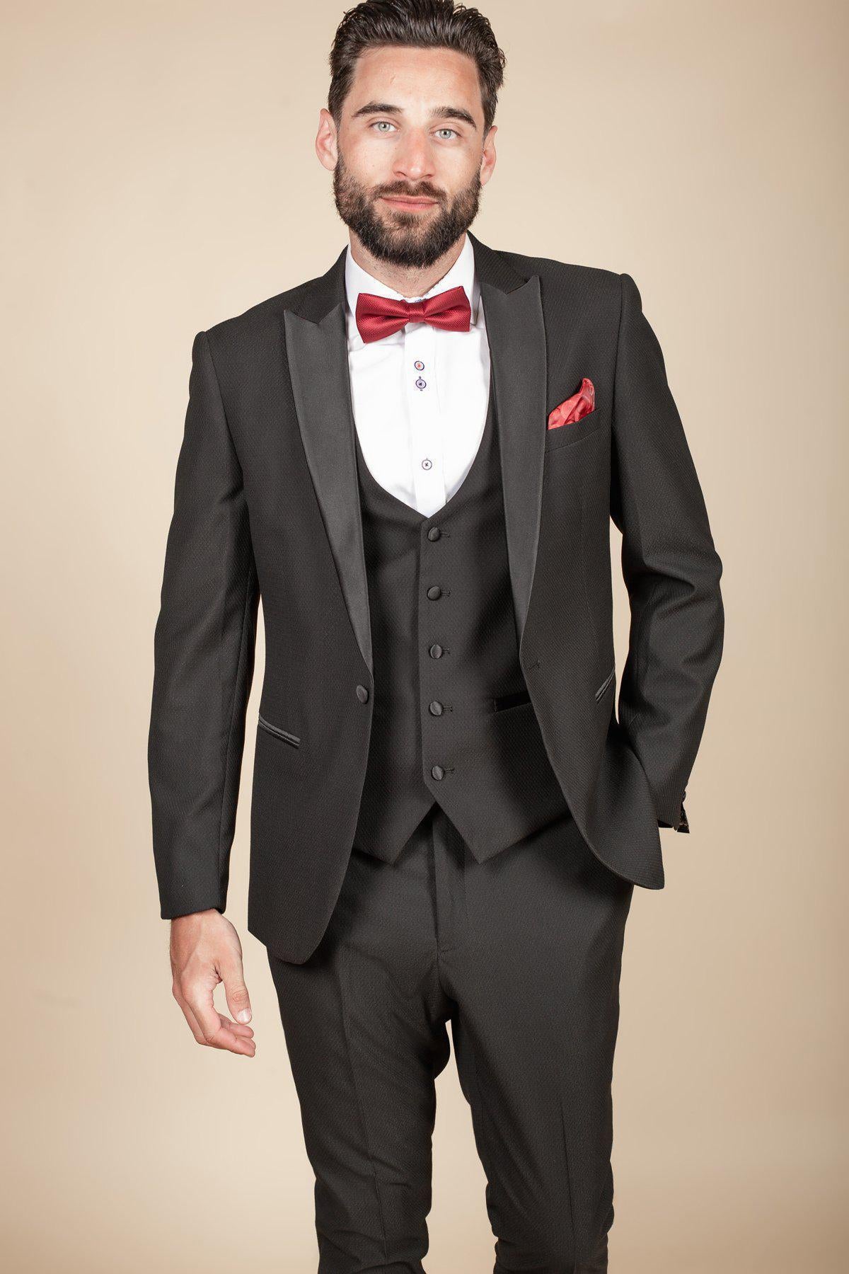 Former Crystal Palace footballer Clinton Morrison wears the marcdarcy DALTON Black Tux Lapel Suit, white shirt, red bow tie, and matching red pocket square, standing against a beige background and looking at the camera with one hand in his pocket.
