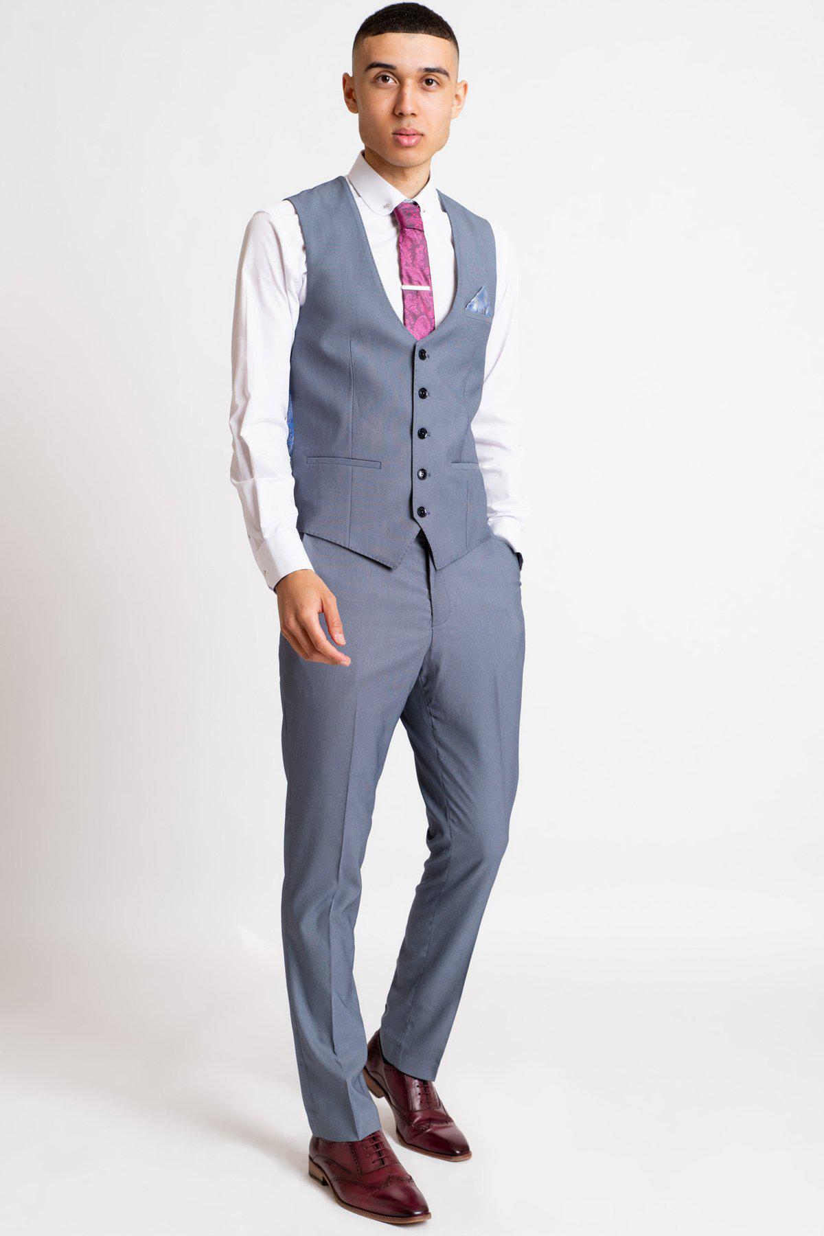 A young man in office attire—a Marc Darcy DANNY blue grey single breasted waistcoat, white shirt, pink tie, and maroon dress shoes—stands with his hands in his pockets against a white background.