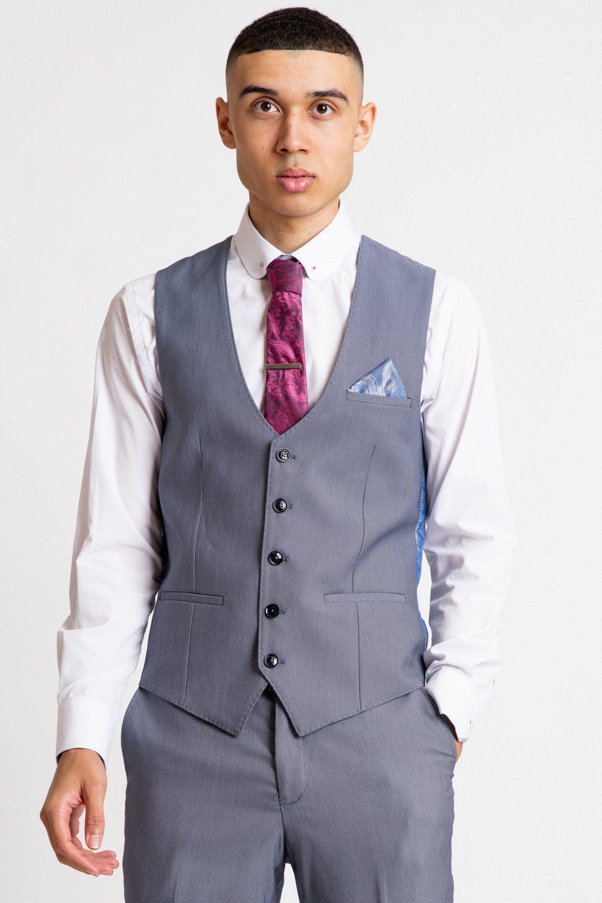 A young man in classic office wear—a Marc Darcy DANNY Blue Grey Single Breasted Waistcoat, grey trousers, white dress shirt, purple tie, and blue pocket square—stands against a plain light background, looking directly at the camera.