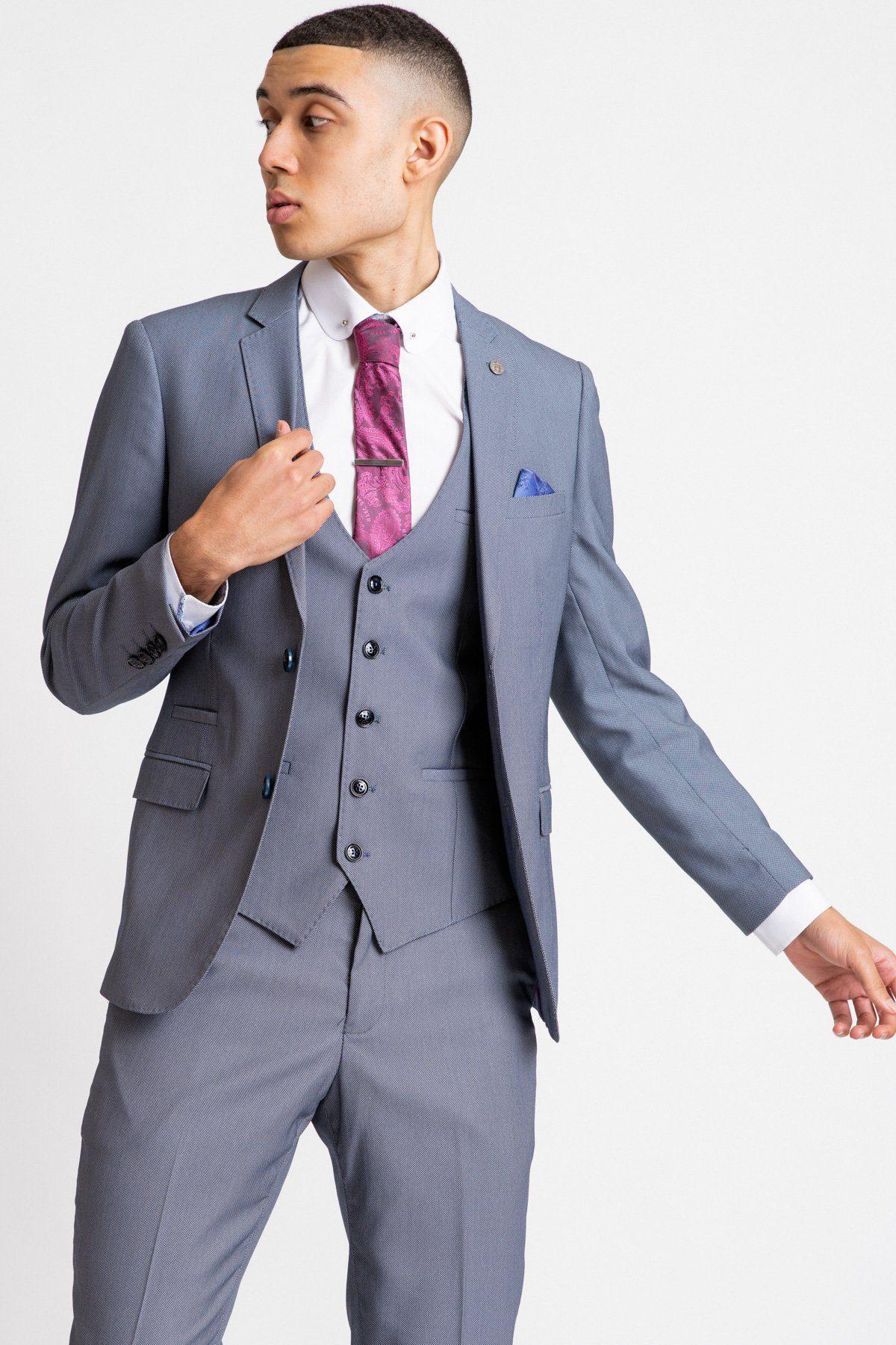 A man in a Marc Darcy DANNY Blue Grey Tailored Blazer three-piece suit with a white shirt and pink patterned tie stands against a white background, looking to the side while adjusting his jacket.