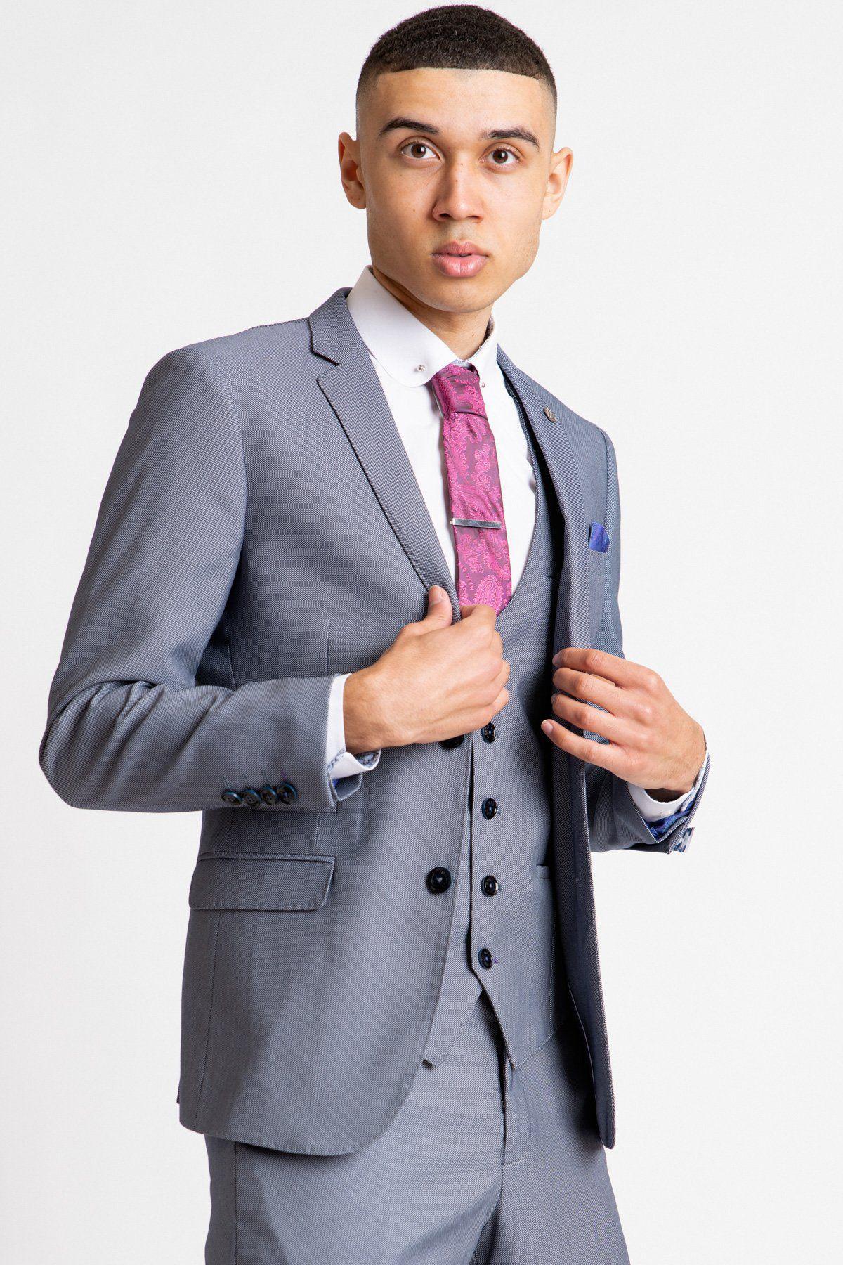 A young man in a Marc Darcy DANNY Blue Grey Tailored Blazer, white shirt, and pink patterned tie stands against a white background, holding his lapels—a contemporary twist on classic workwear style.